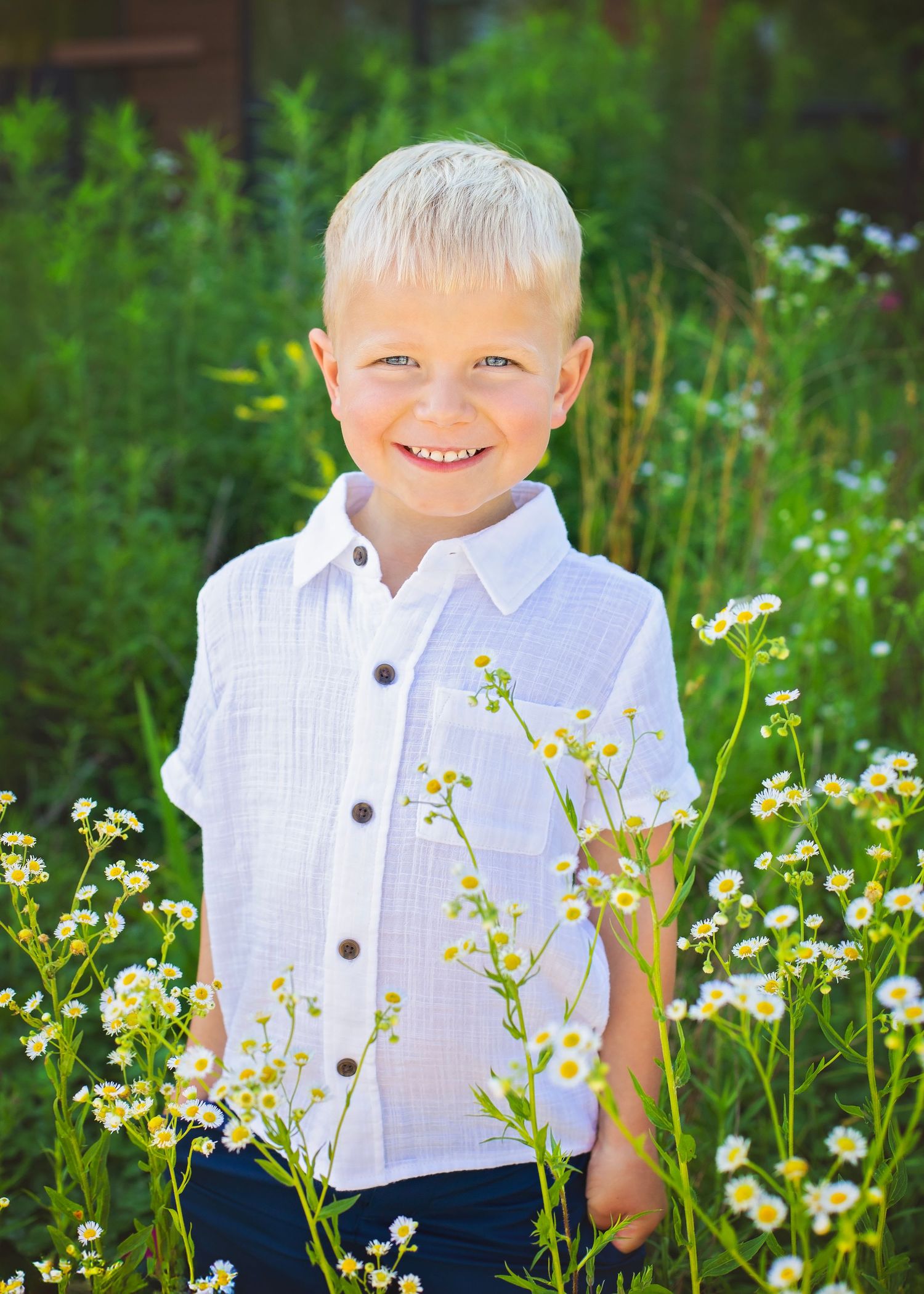 Happy blonde boy in a white shirt smiles while standing among blooming white wildflowers outdoors.