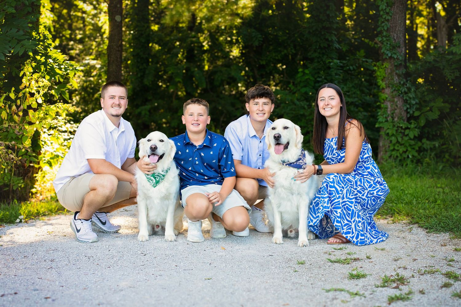 Four siblings kneeling with two fluffy white dogs on a sunny gravel path surrounded by trees.