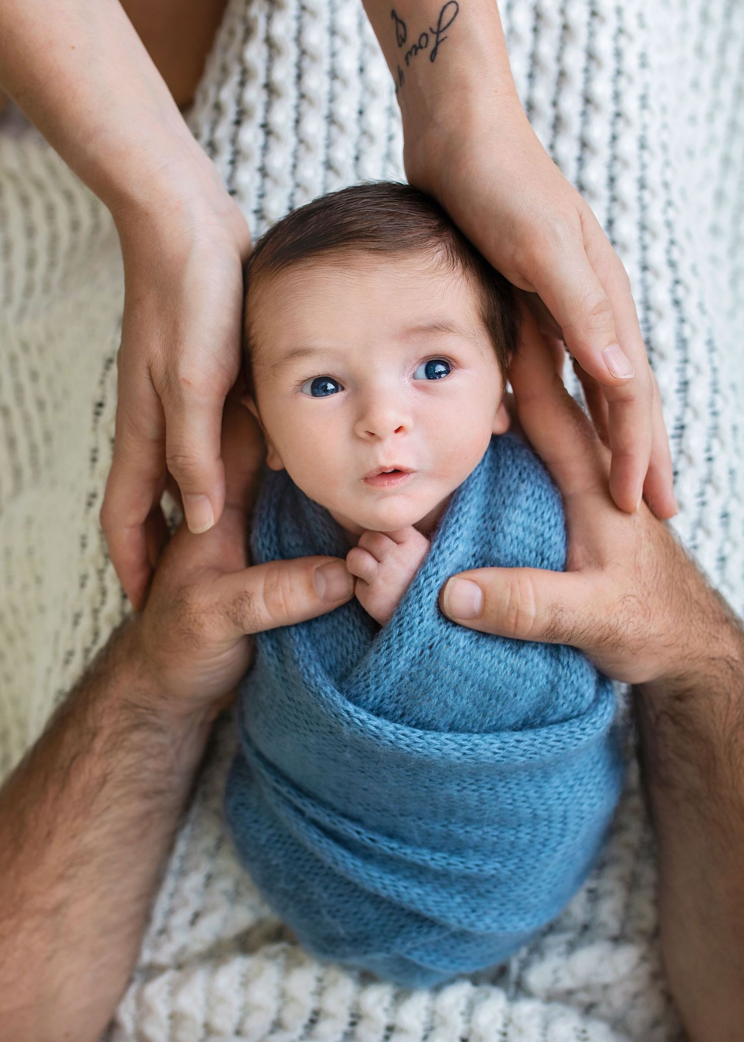 Close-up of newborn baby boy swaddled in blue knit wrap with parents hands gently supporting him.