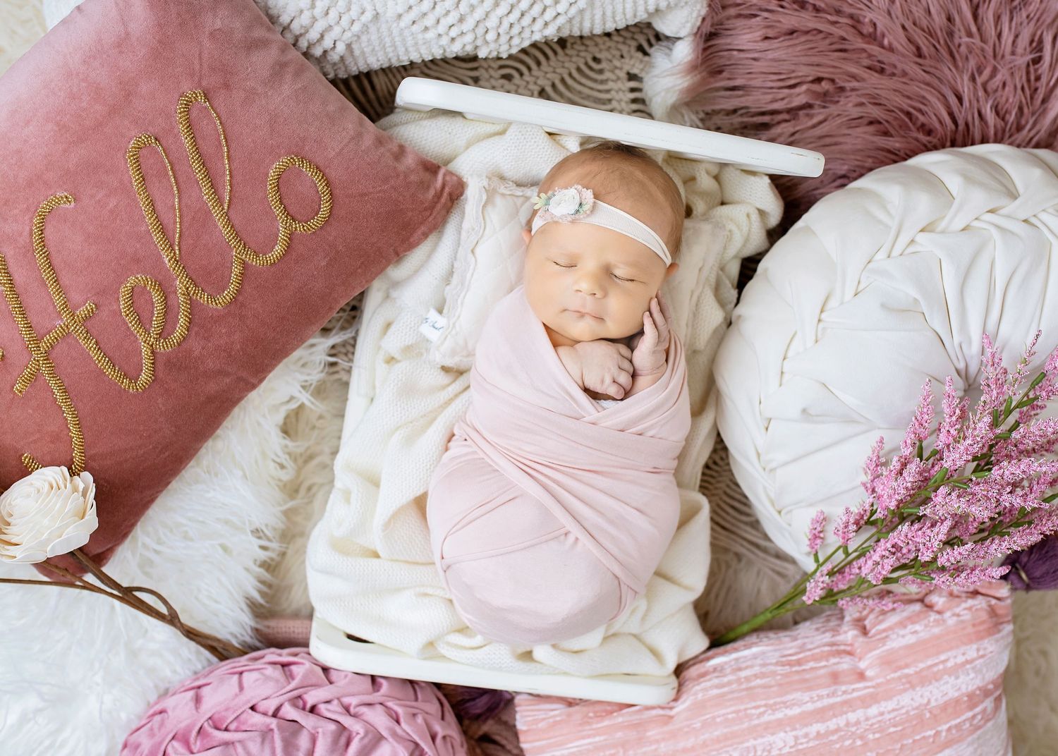 Newborn baby girl swaddled in pink wrap sleeping in white mini crib surrounded by pink pillows and flowers.
