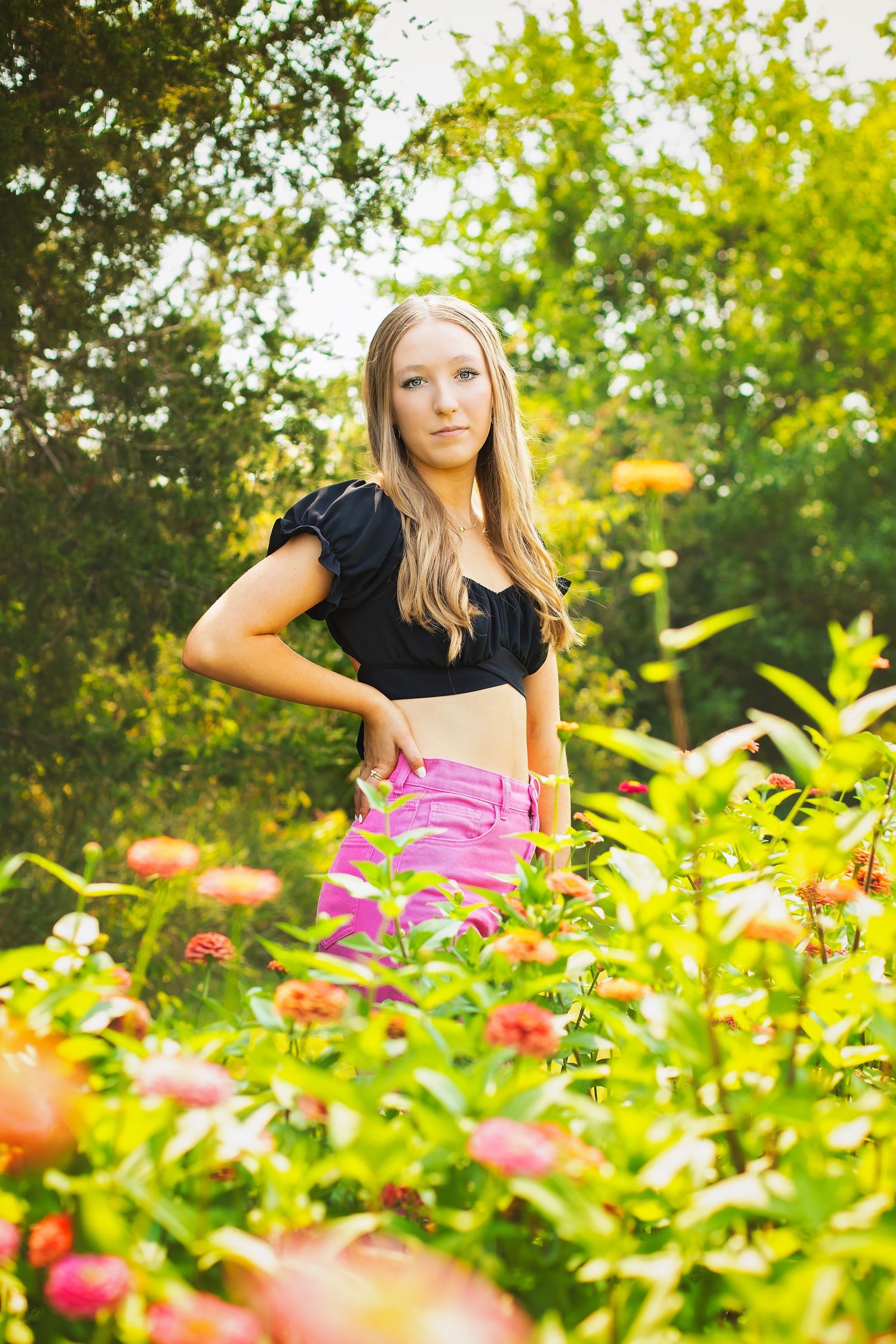 Blonde teenage girl in black crop top and pink jeans standing among vibrant garden flowers.