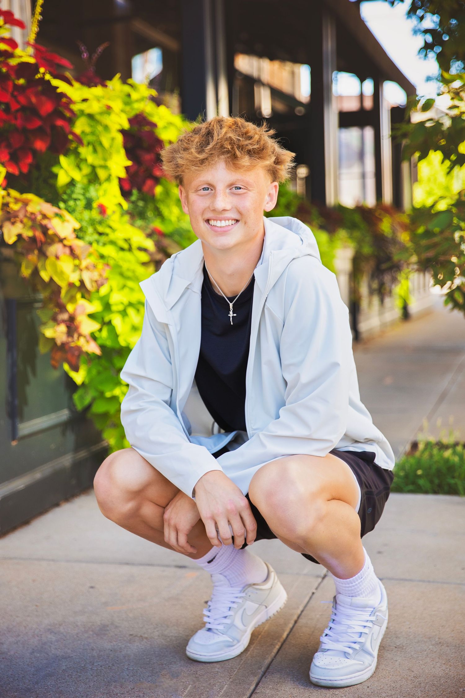 Smiling teen boy with curly blonde hair crouches on sidewalk wearing white hoodie near colorful flowers.