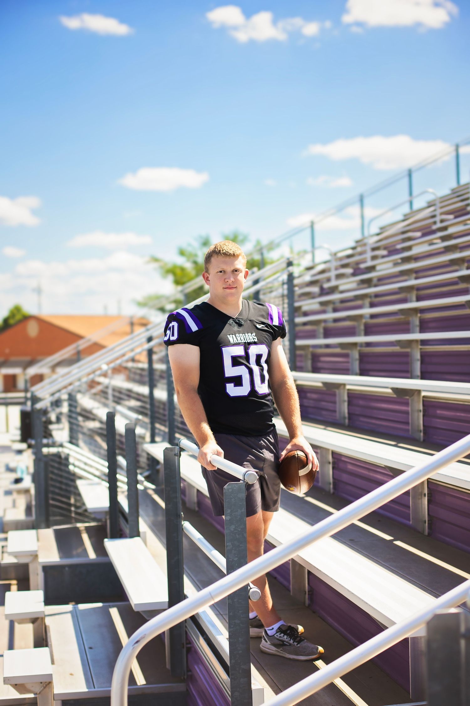 Football player wearing number 50 Warriors jersey holds helmet in stadium bleachers.
