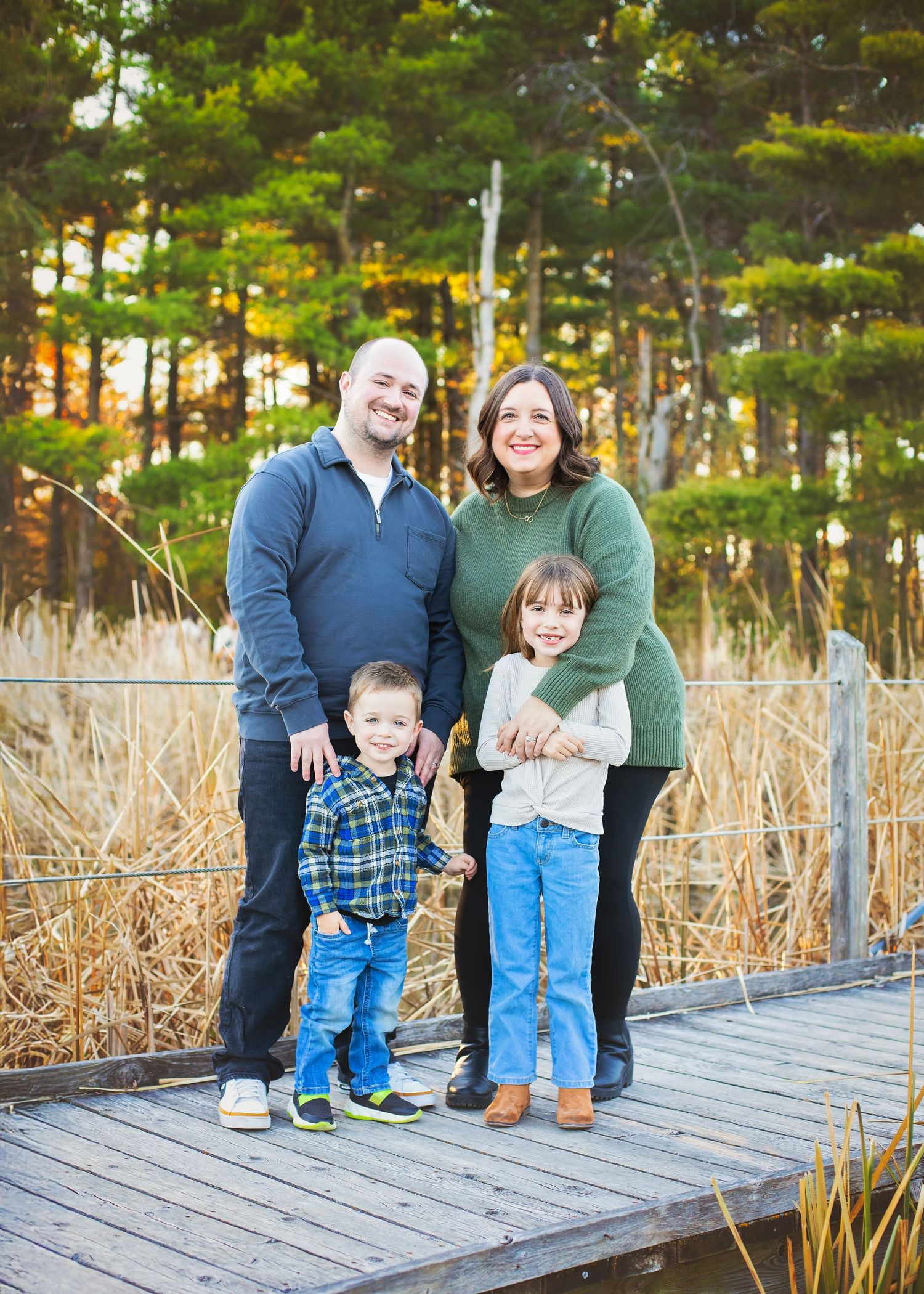 Happy family of four standing on wooden boardwalk surrounded by autumn trees and golden marsh grass.