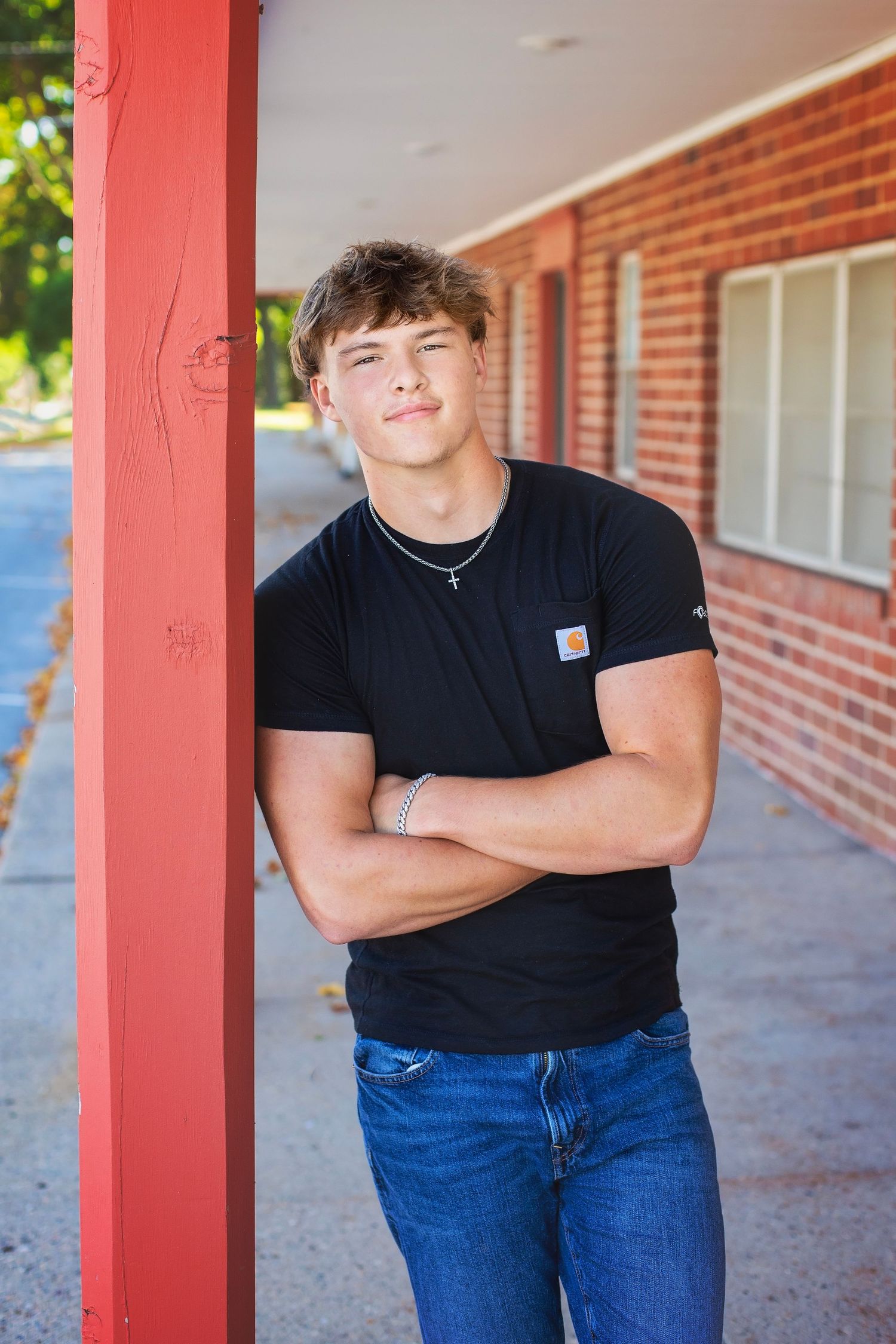 Athletic teen in black Carhartt t-shirt leans against red brick pillar with arms crossed.