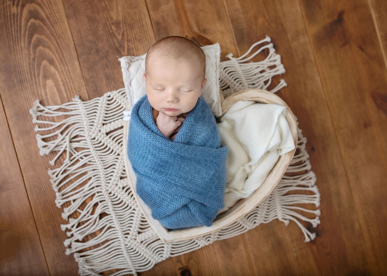 Sleeping newborn baby wrapped in blue knit swaddle in a round basket on macrame mat.