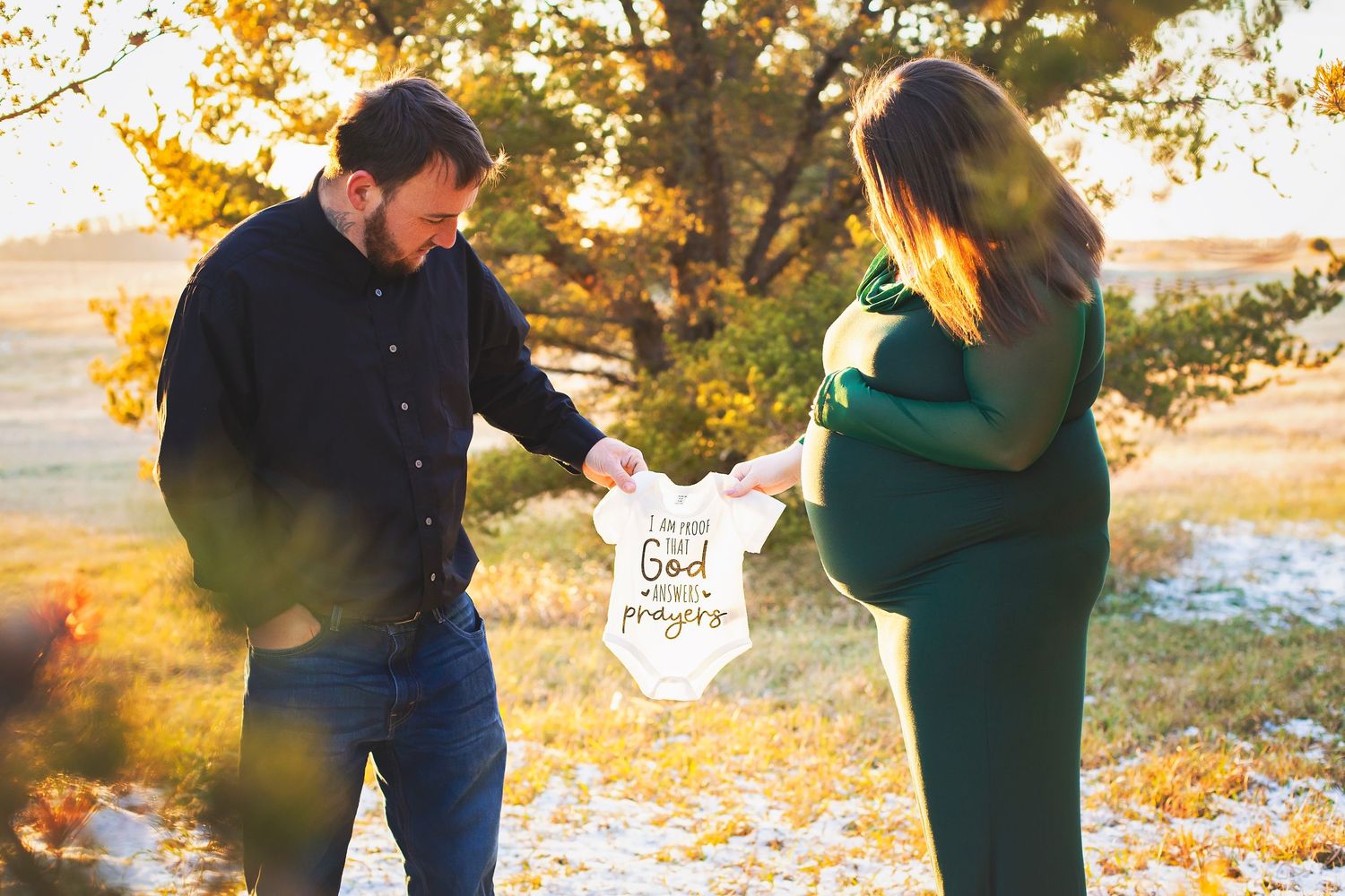 Couple holding a baby onesie during outdoor maternity photoshoot in autumn setting.