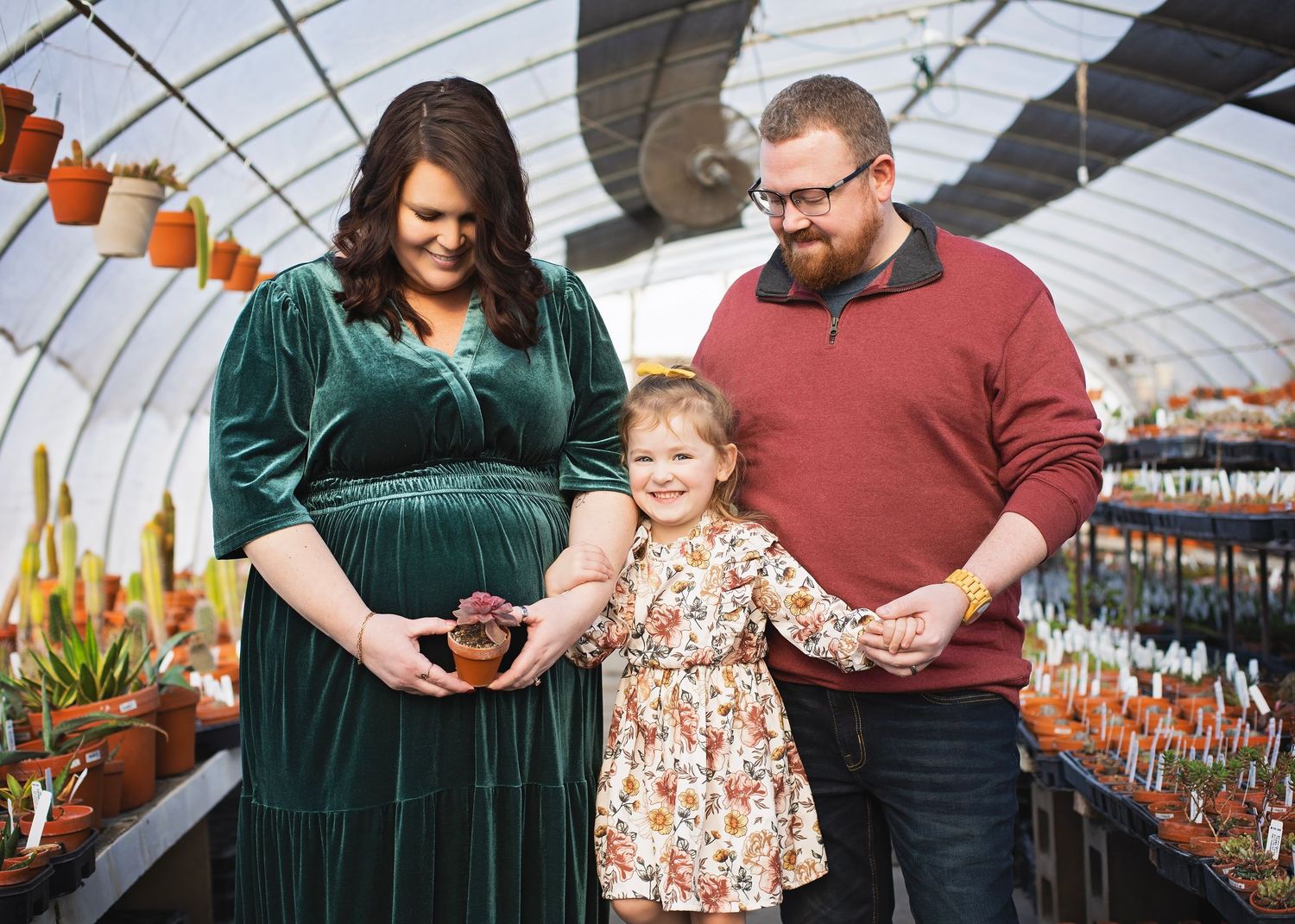 Smiling pregnant woman in green dress and bearded husband walk with toddler daughter through greenhouse.