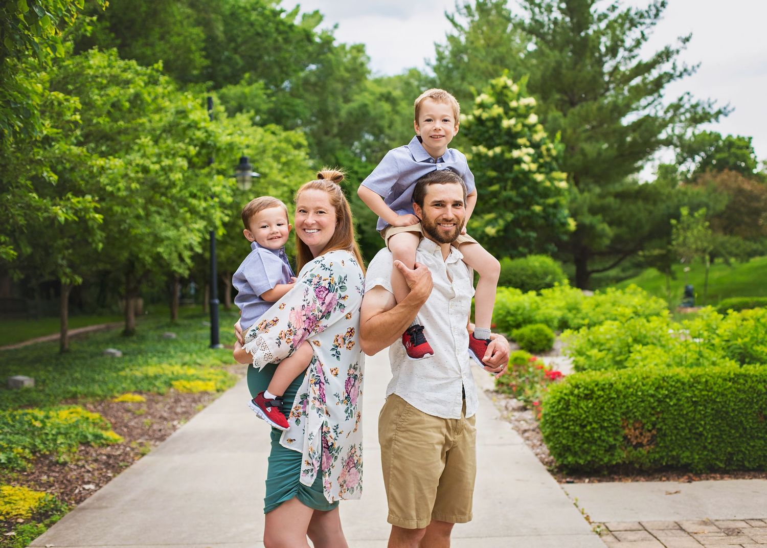 Smiling family of four enjoys outdoor walk, with father carrying son on shoulders and mother holding toddler.