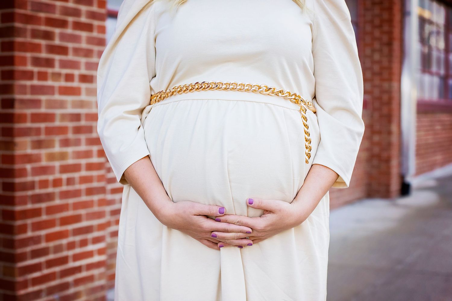 Close-up of pregnant woman in white dress with gold chain belt holding baby bump.