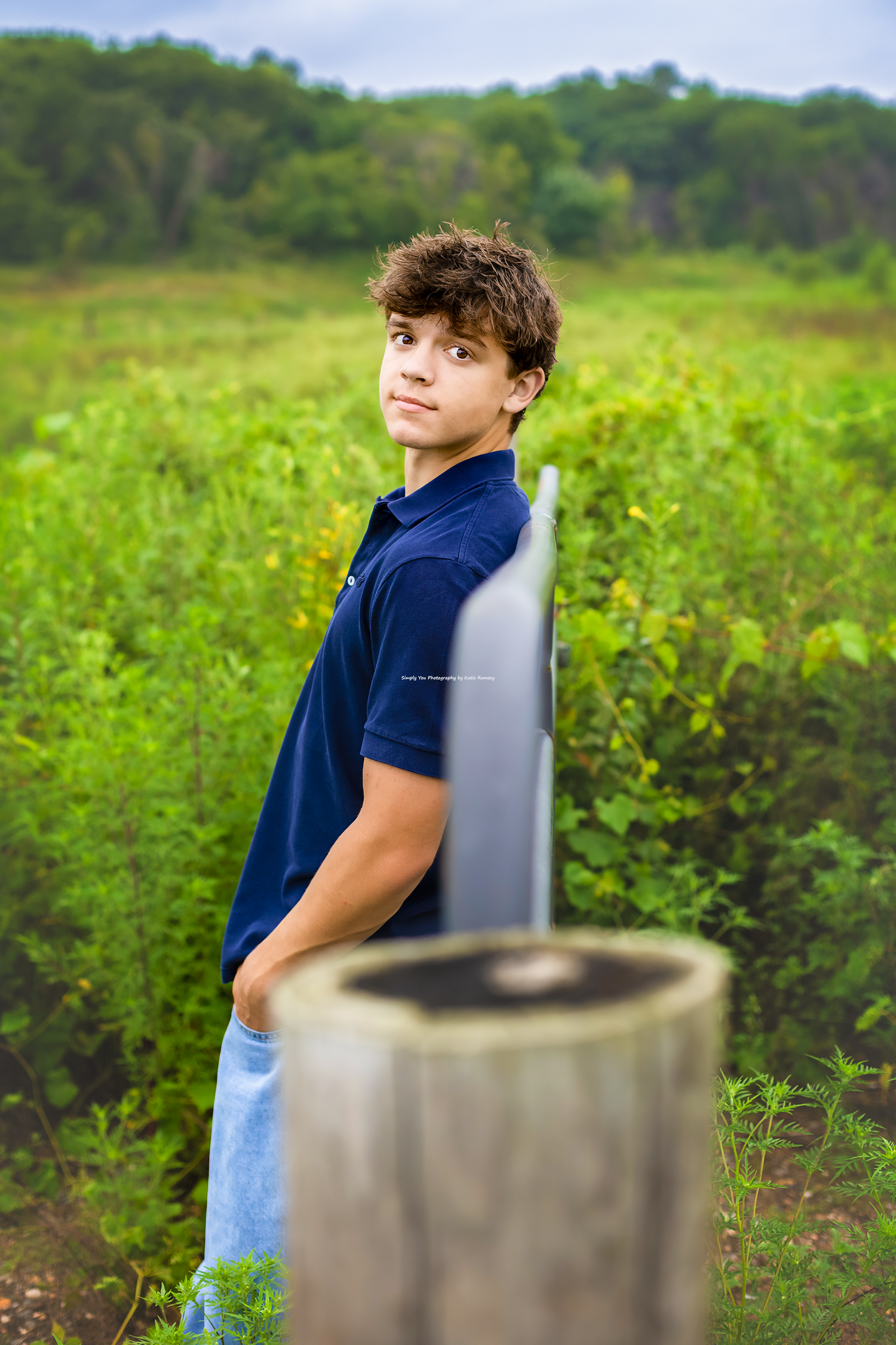 Teen boy in navy polo shirt posing outdoors in lush green field near wooden fence post.