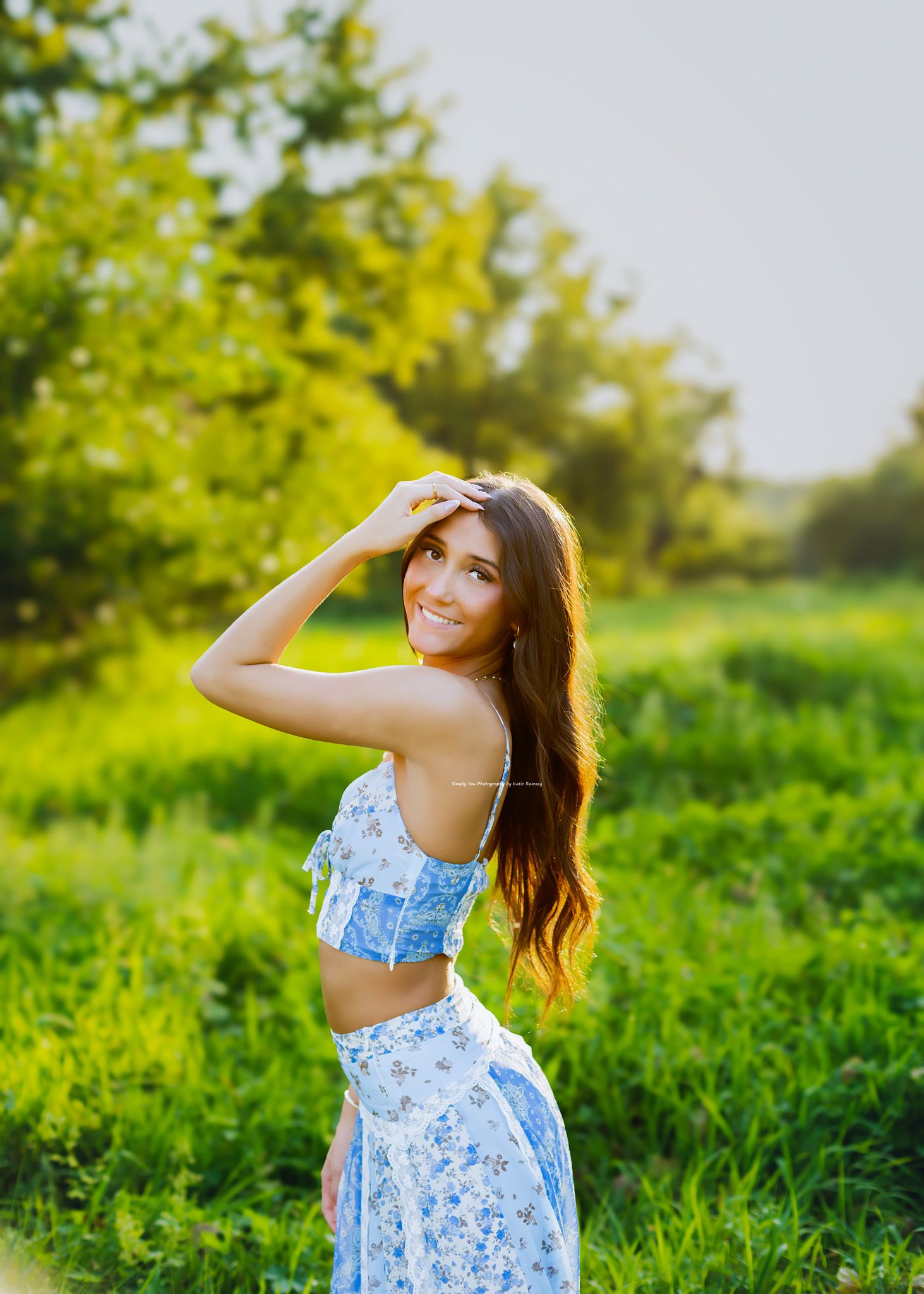 Young woman in blue floral crop top and skirt smiling outdoors in a lush green field during golden hour.