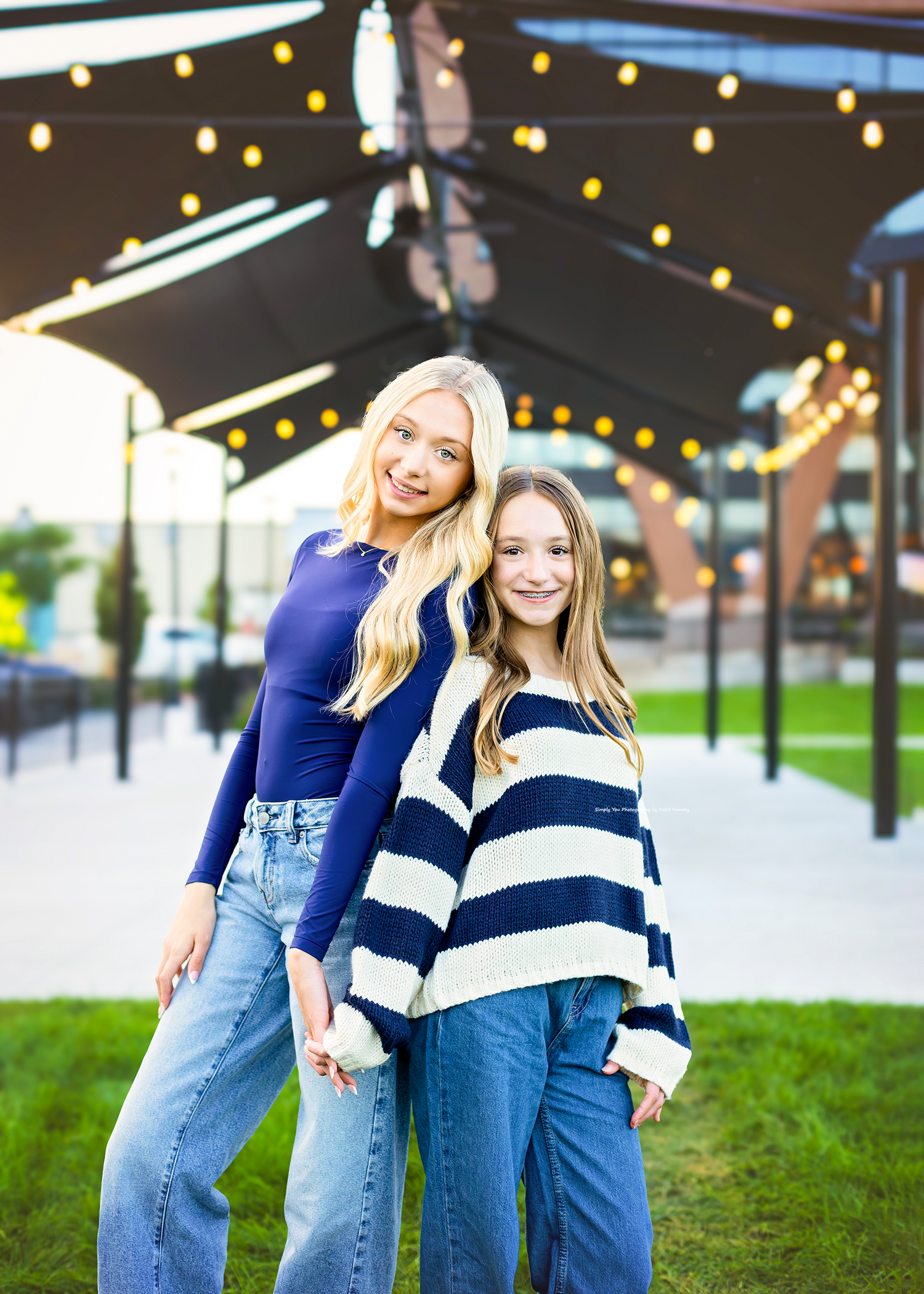 Two young blonde girls pose outdoors in blue outfits, one in a navy top and jeans, the other in a striped sweater.