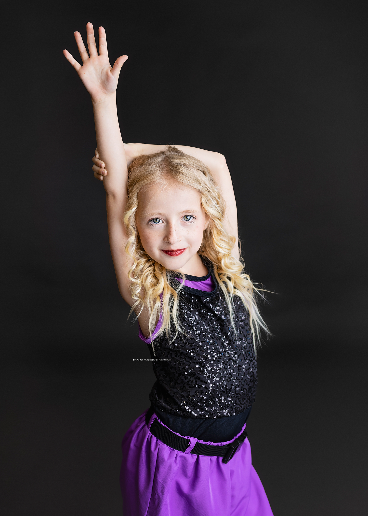 Young blonde girl in black sequin top and purple skirt poses with arm raised in a dance studio portrait.