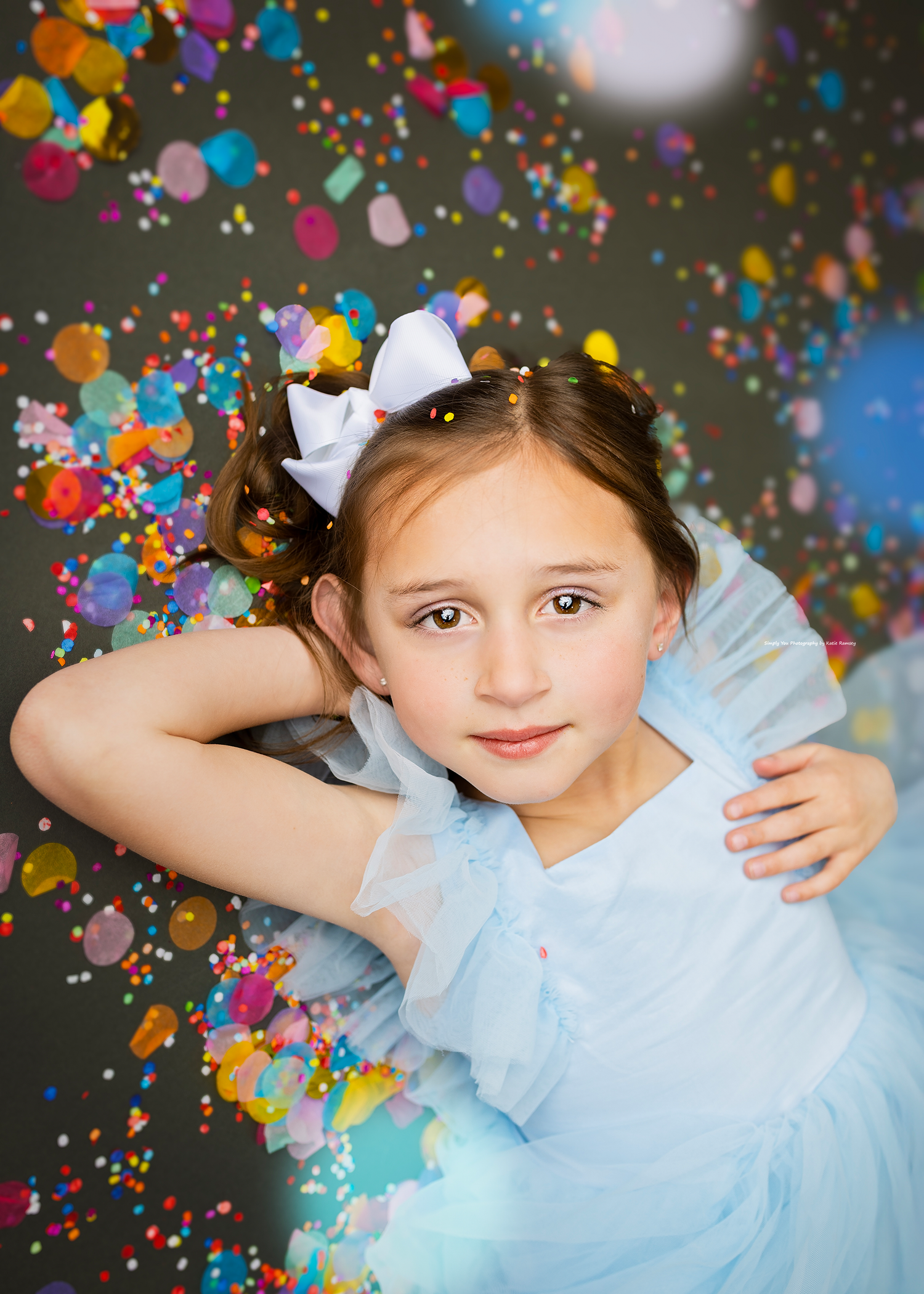 Young girl in blue tulle dress with white bow lying among colorful confetti.