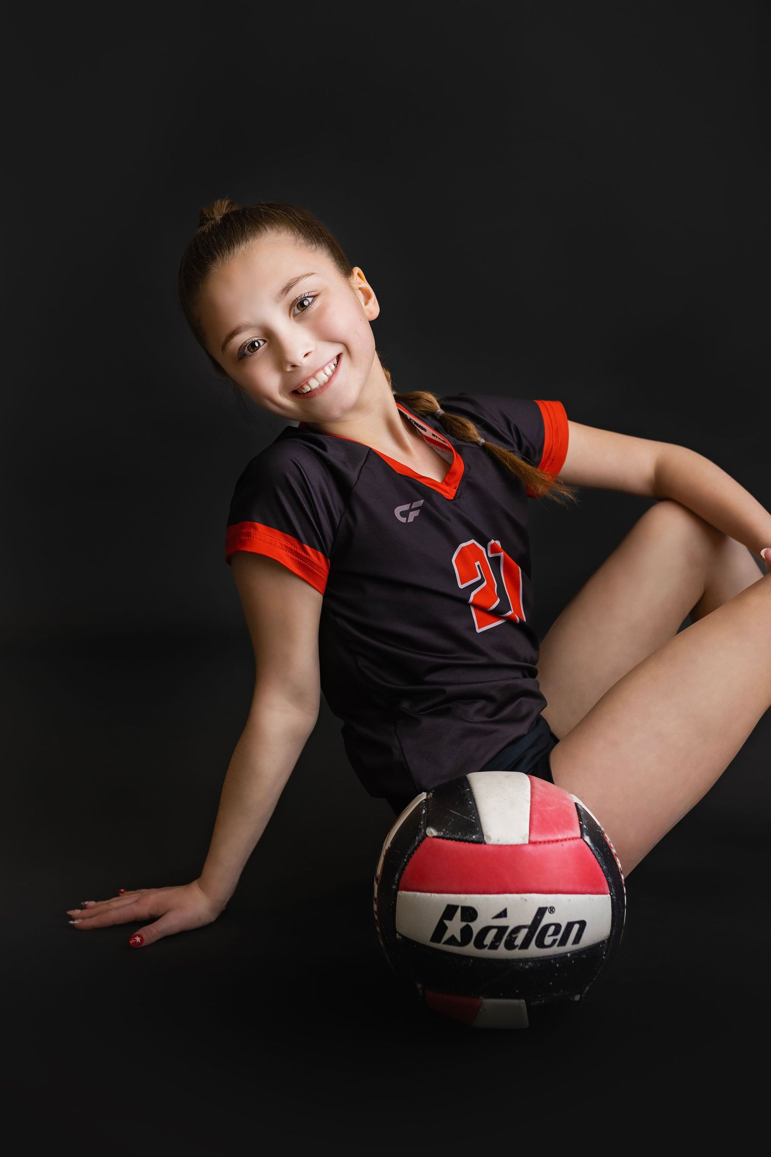 Smiling young girl in black number 21 volleyball jersey poses with a Baden volleyball on a dark studio background.