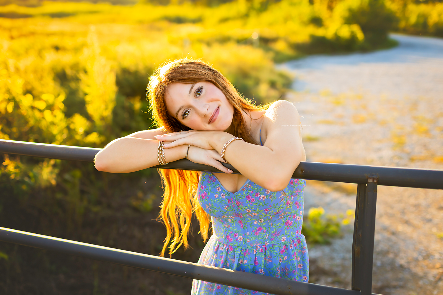 Young woman with long red hair in floral dress leaning on metal railing in golden sunlight outdoors.