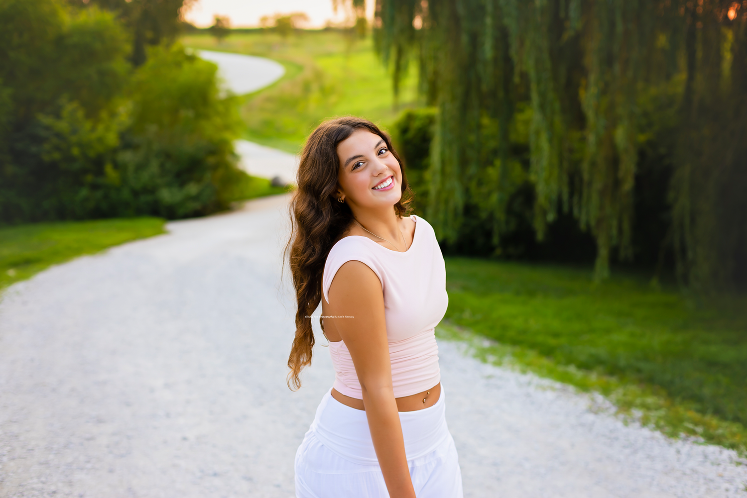 Young woman in white outfit smiling over shoulder on scenic gravel path surrounded by lush green trees.