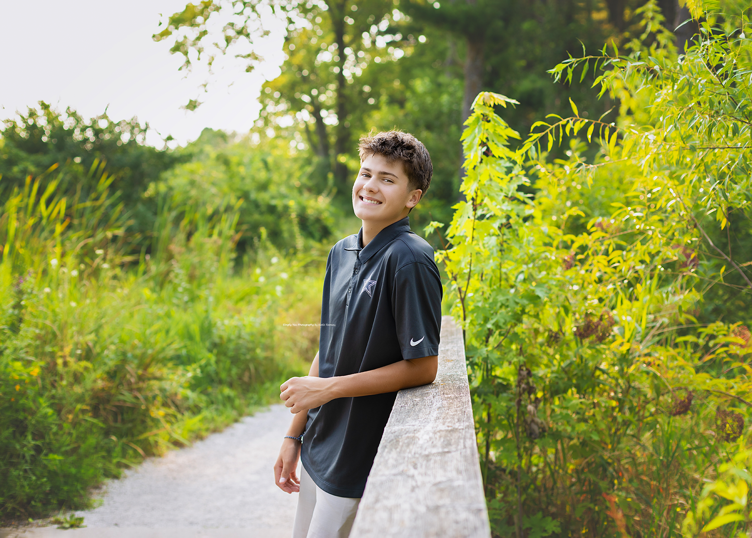 Smiling teenage boy in navy polo shirt leaning on stone wall surrounded by lush green trees on a nature path.