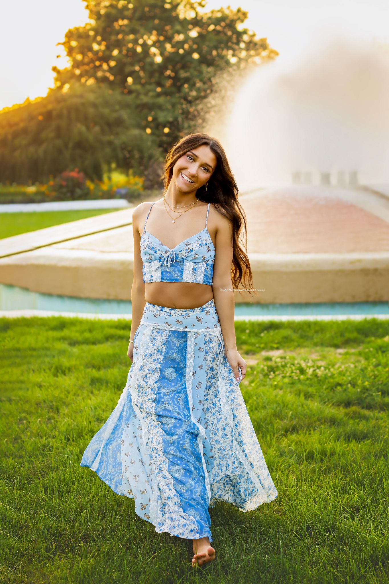 Smiling young woman in blue floral crop top and maxi skirt walking near a fountain at sunset.