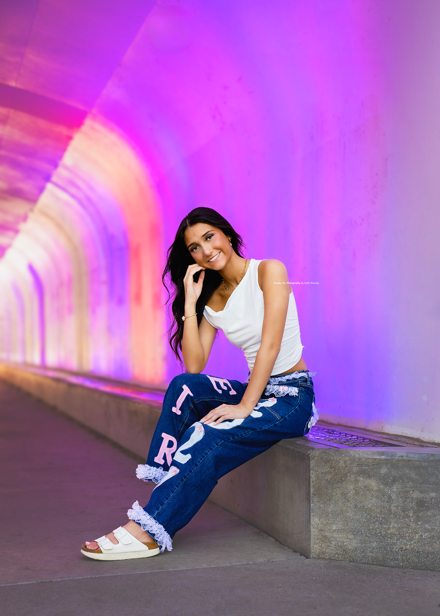 Young woman in white tank top and decorated jeans sits on concrete bench in colorful illuminated tunnel.