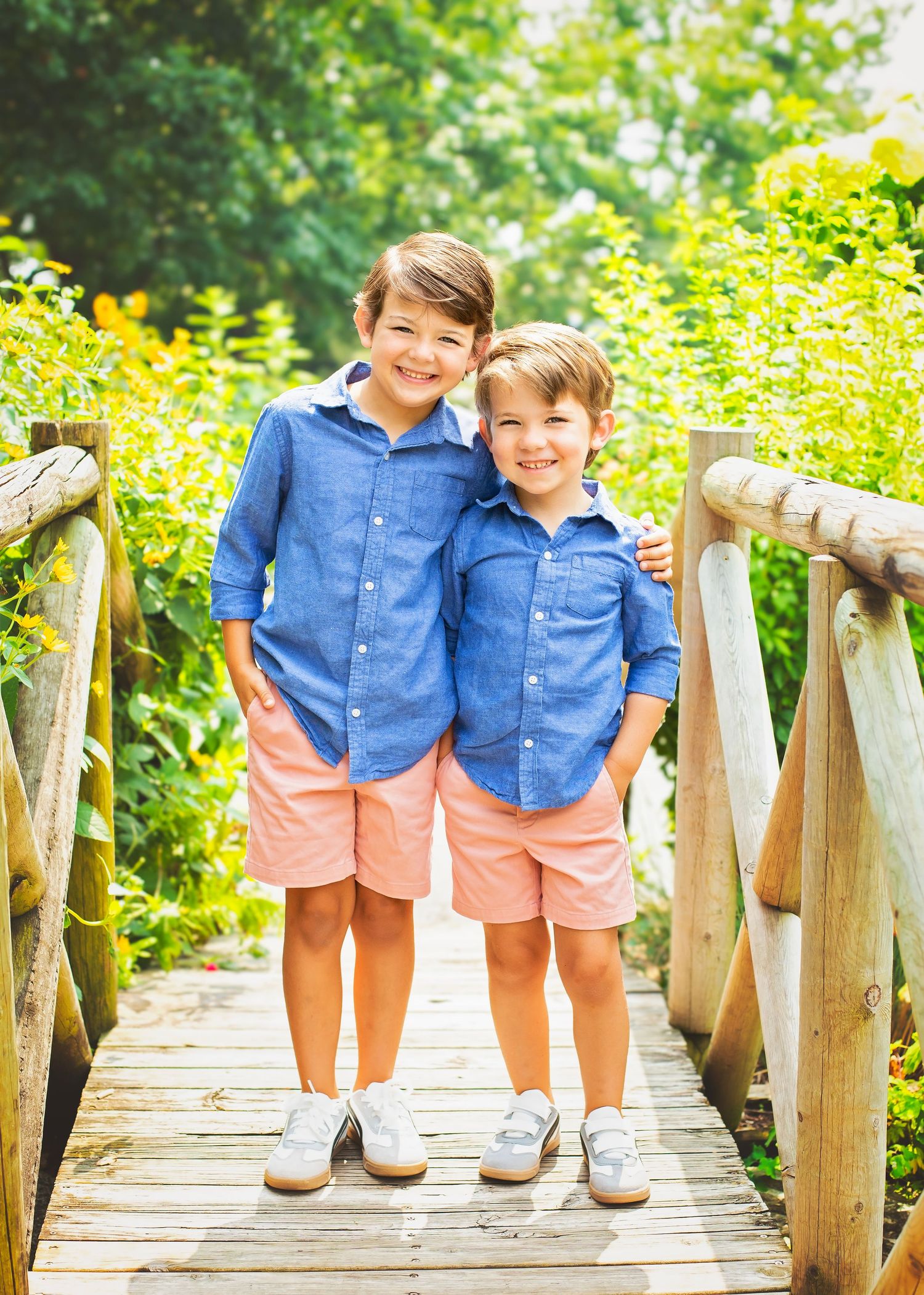 Two brothers in matching blue shirts and pink shorts smile together on a wooden garden bridge.