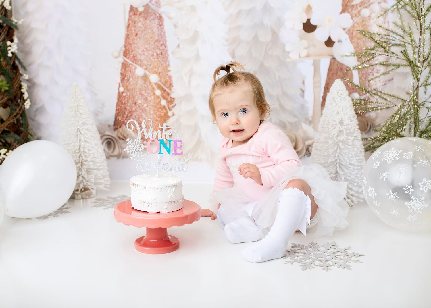 Baby girl in pink outfit sitting near a white cake with a Winter ONEderland topper at her first birthday.