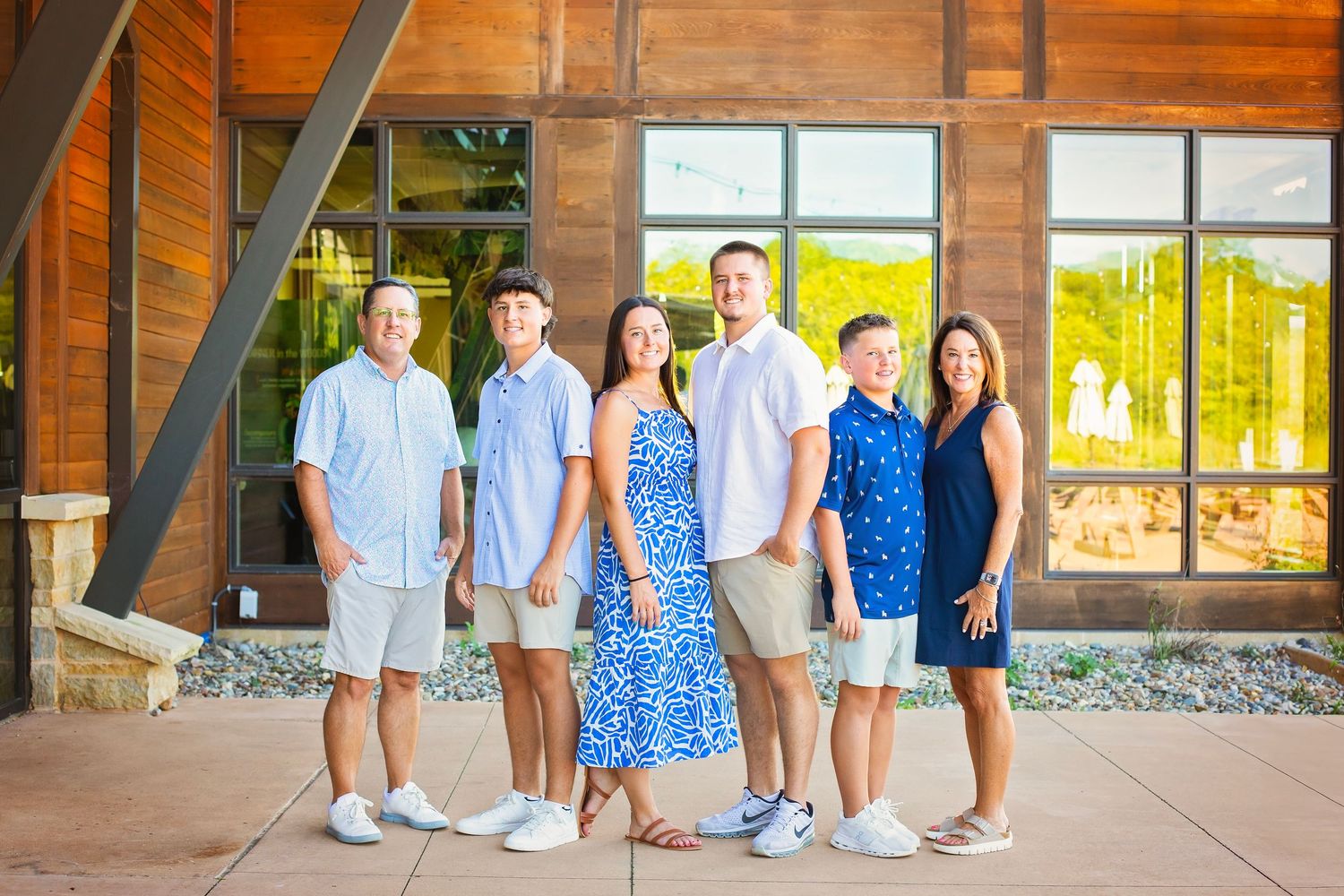 Family of six posing and smiling together in front of a modern wooden building on a sunny day.