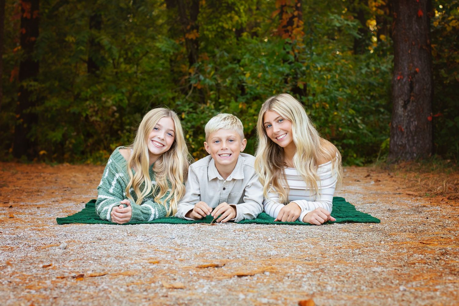 Three blonde siblings smile while lying on a green blanket on a leaf-covered forest path in fall.