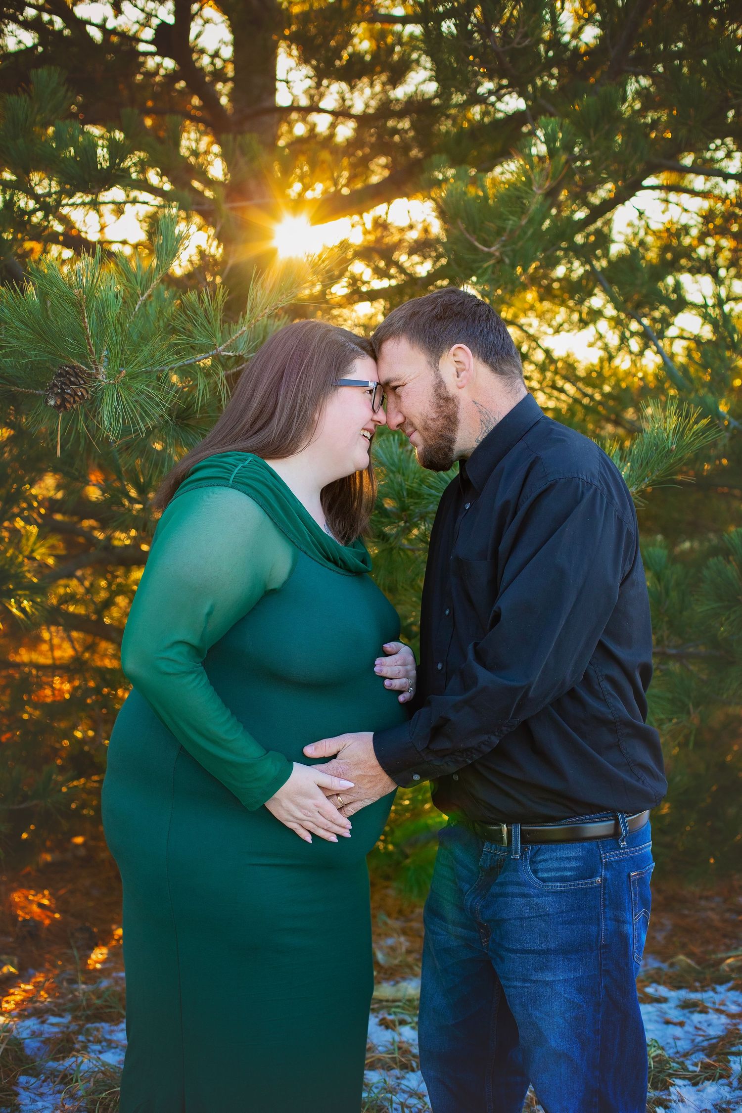 Pregnant woman in green dress and bearded partner touching foreheads at sunset in pine forest.