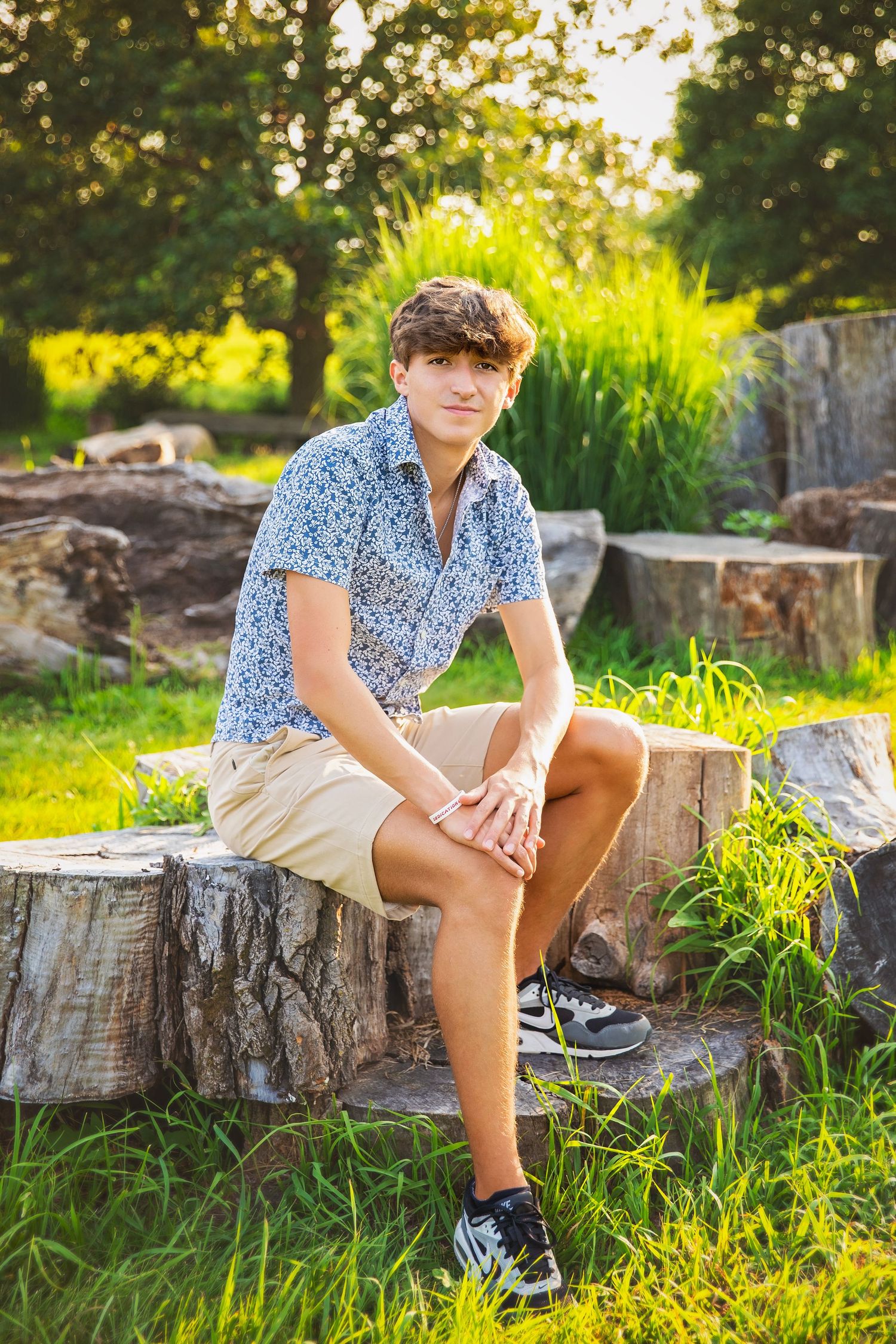 Teen boy in blue floral shirt and khaki shorts sitting on a tree stump in a sunny green park.