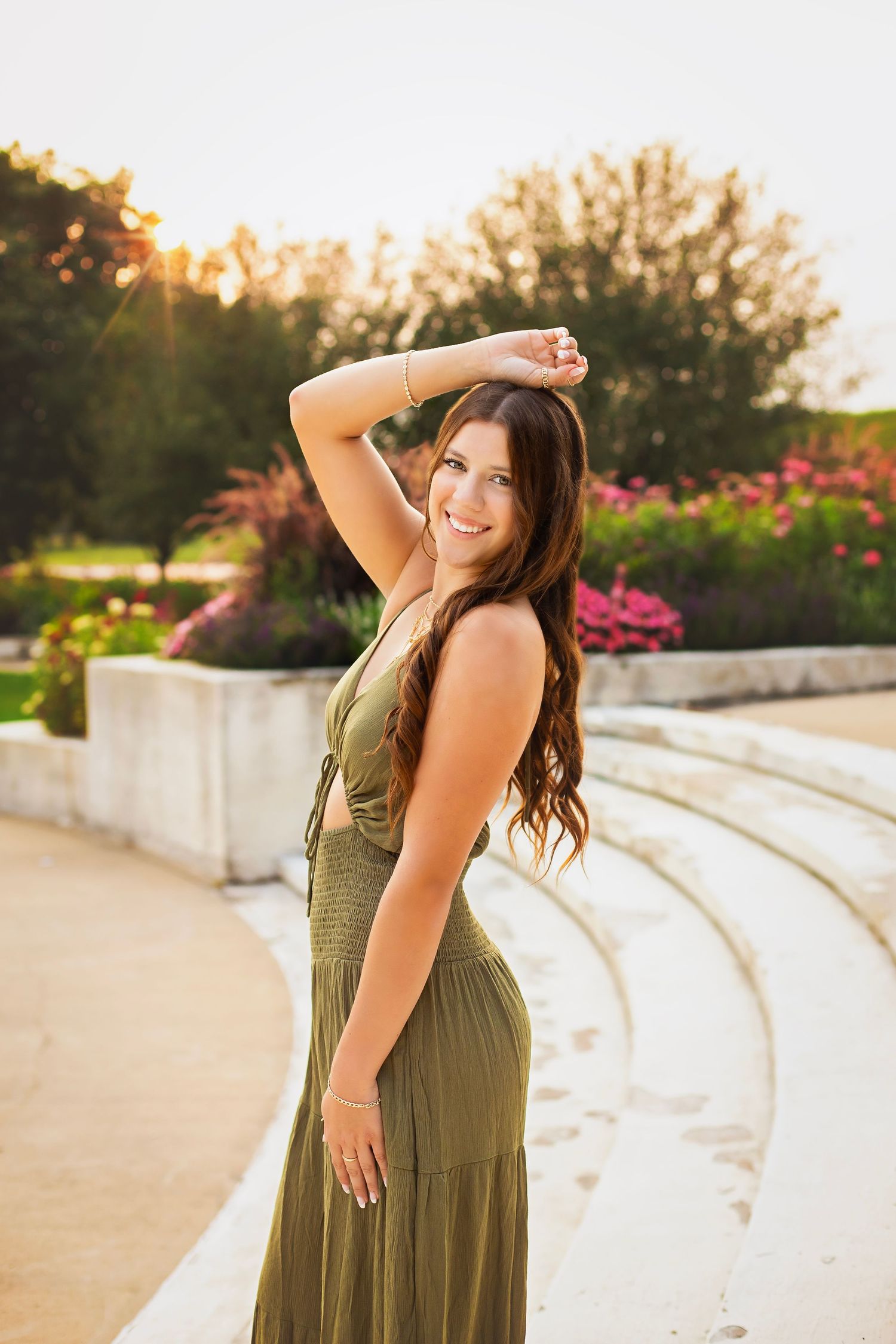 Smiling young woman in olive dress poses at golden hour near garden amphitheater.