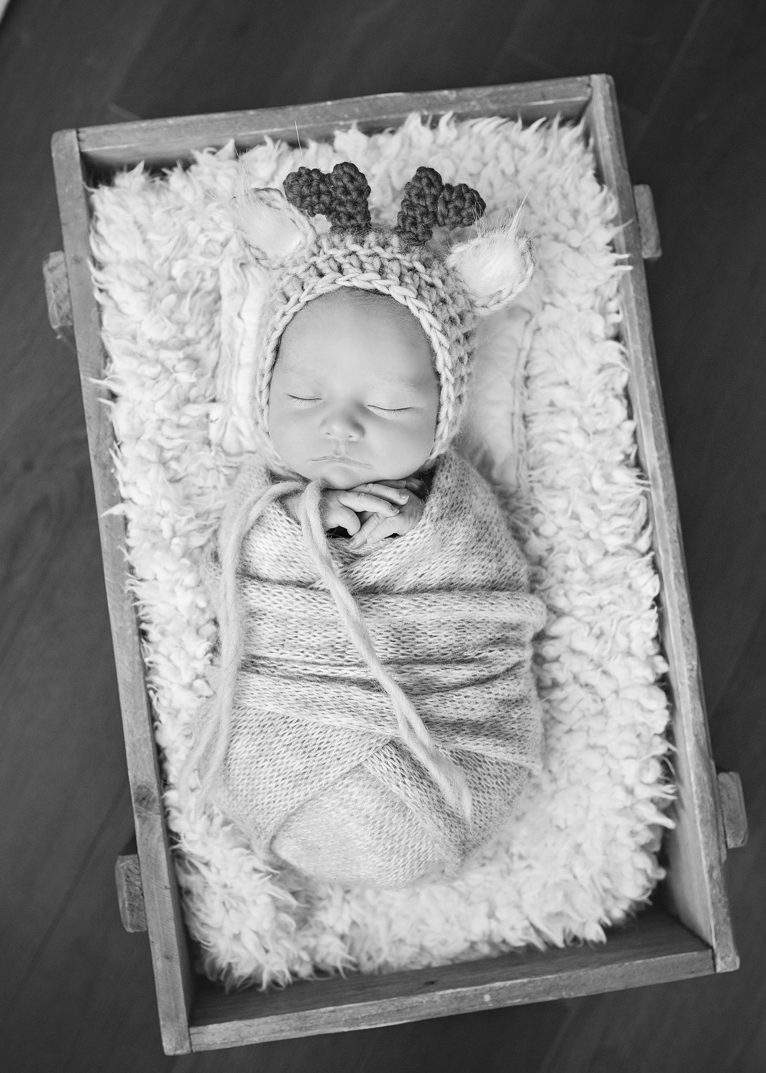 Sleeping newborn baby swaddled in a reindeer bonnet inside a rustic wooden crate with fluffy white lining.