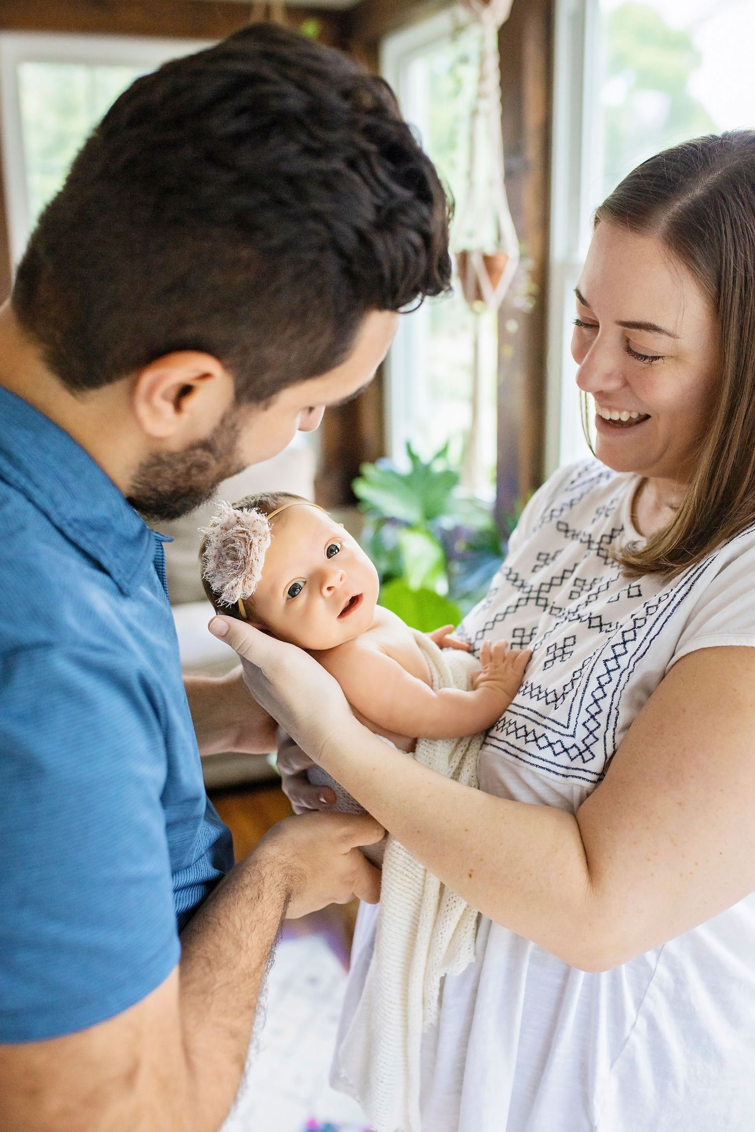 Joyful parents smiling and holding their alert newborn baby girl with floral headband indoors.