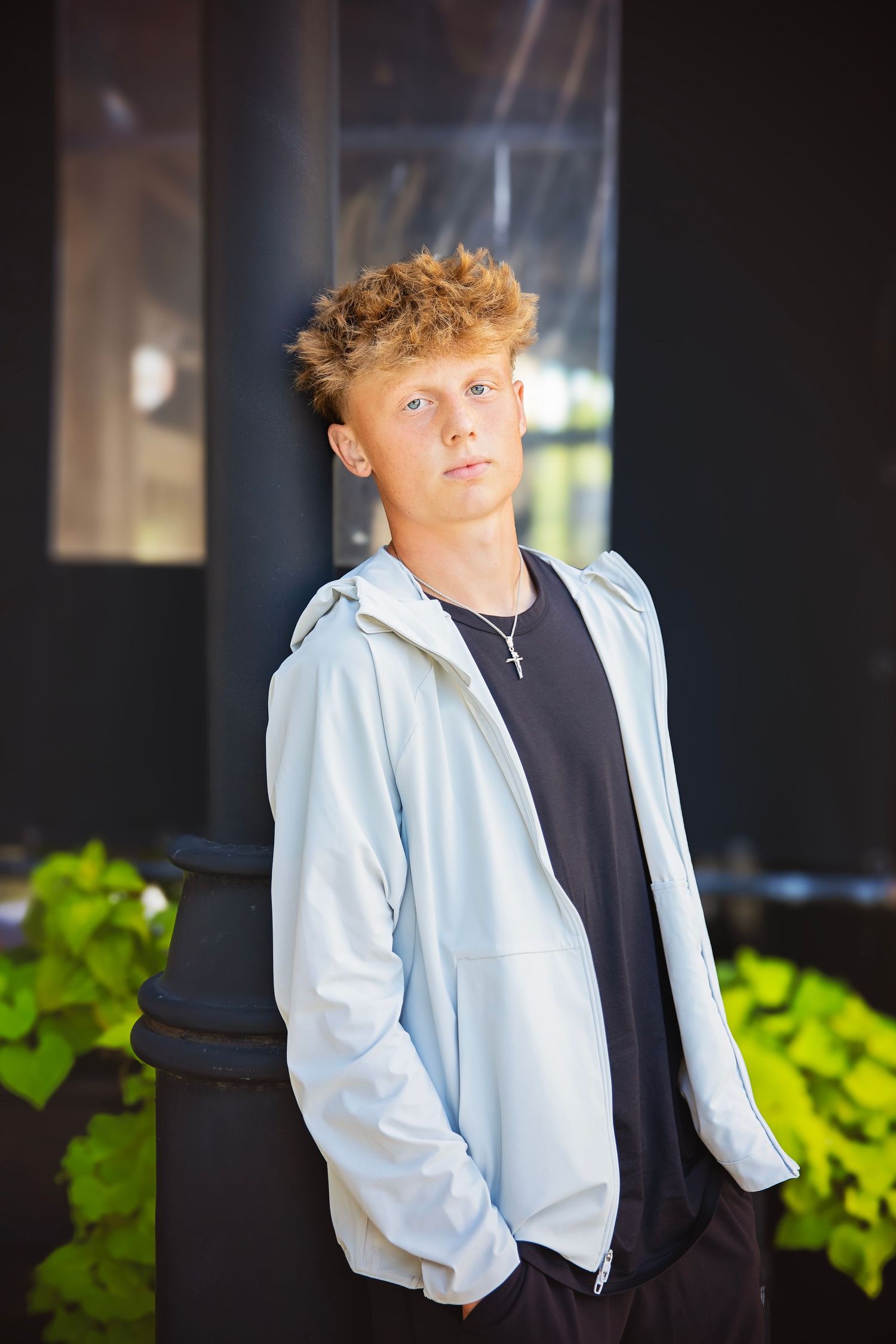 Teen boy in light blue zip hoodie and black shirt standing near dark pillar with green plants.