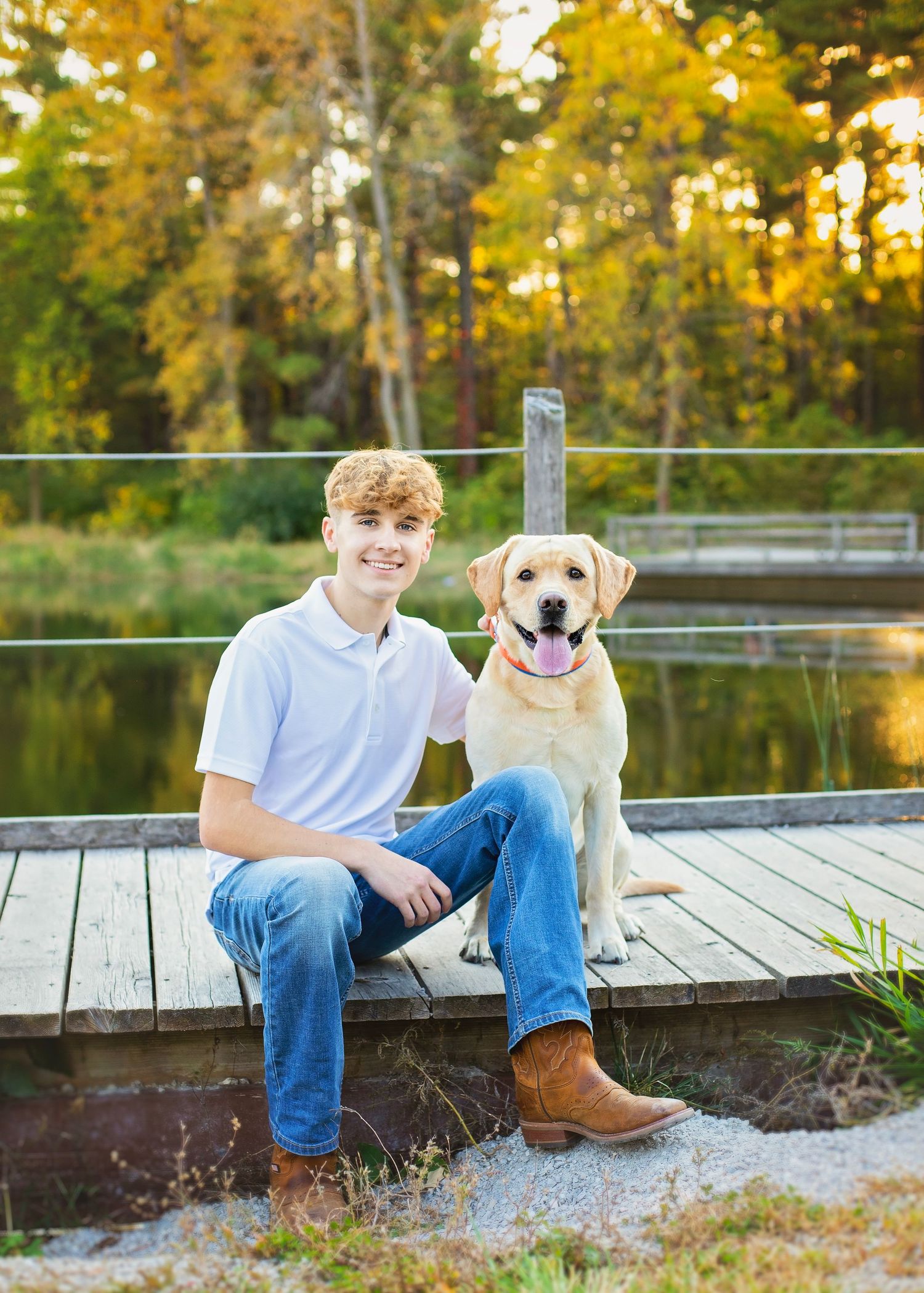 Smiling teen boy sitting on a dock with a yellow Labrador Retriever during fall senior photo session.