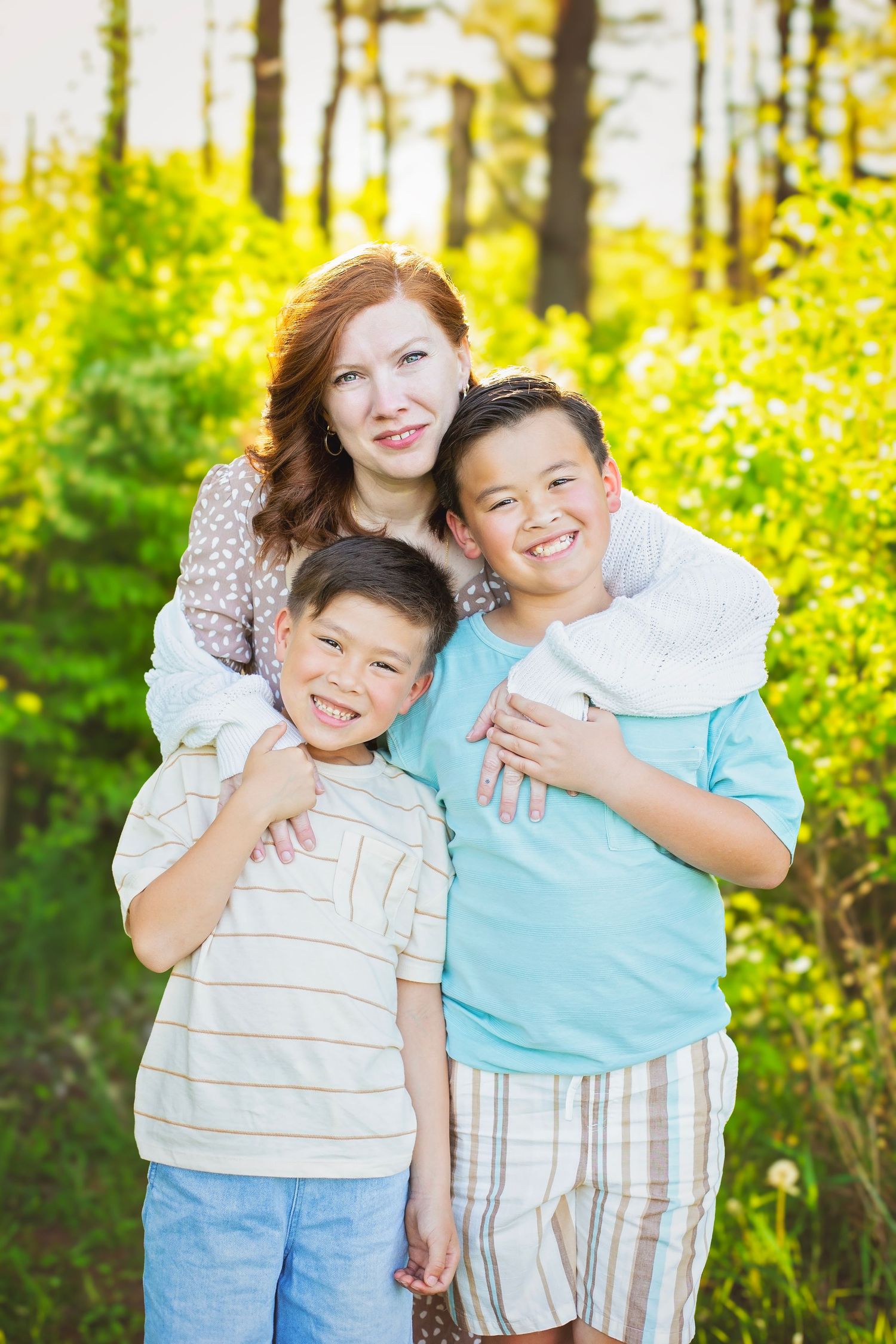 Red-haired mother hugs two smiling boys in a sunny green forest setting.