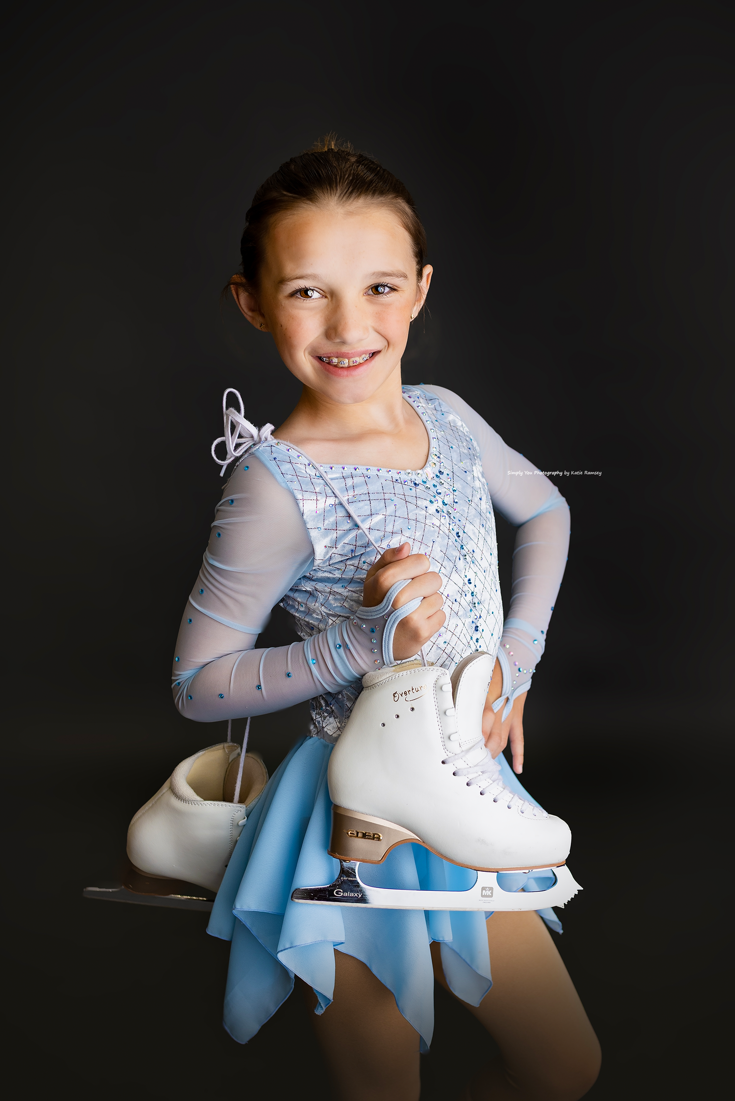 Young girl in light blue figure skating dress holding white ice skates, smiling against dark background.