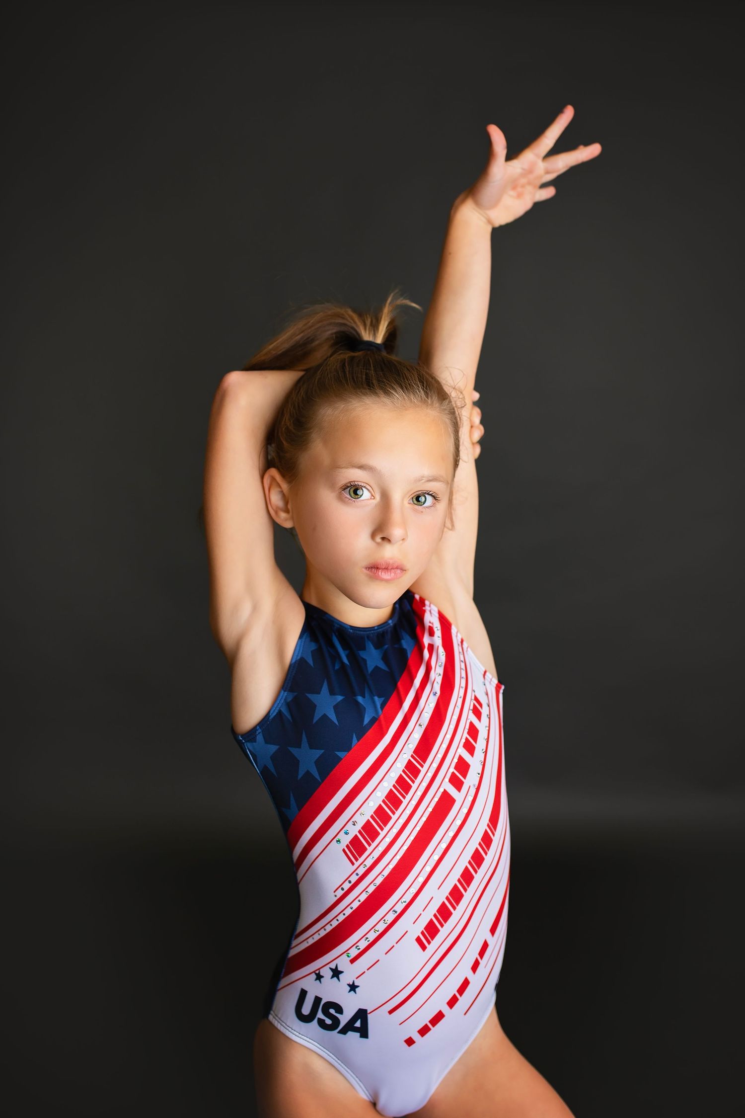 Young gymnast in USA leotard raises arms overhead in an elegant pose.