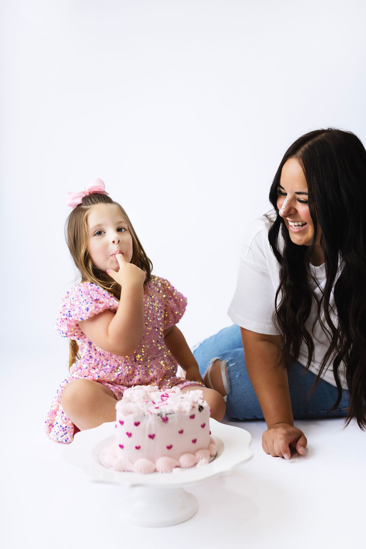 Little girl in pink sequin dress tastes cake frosting while mom in white top laughs beside pink birthday cake.