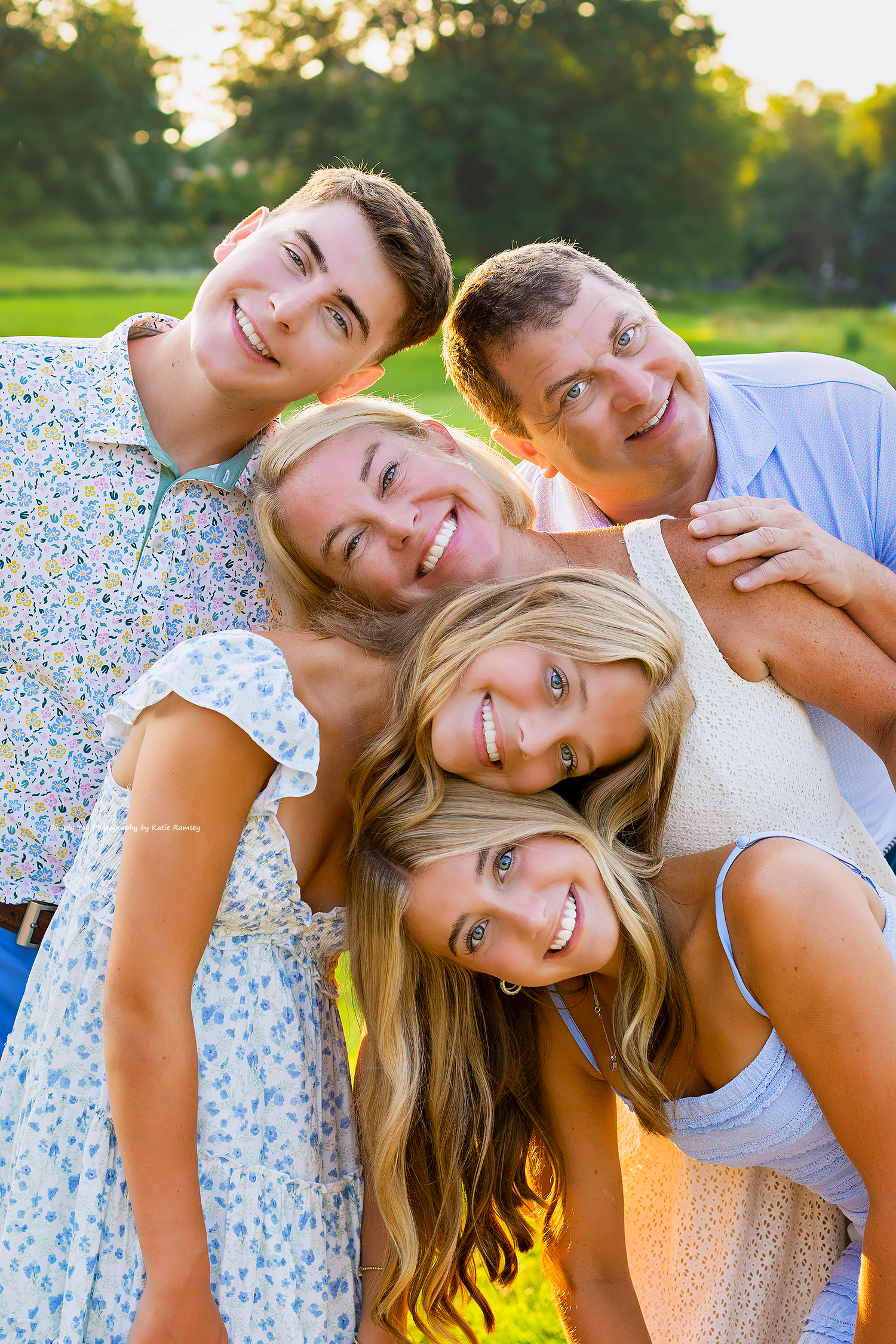 Happy family of five smiling together outdoors in a sunny park, wearing casual summer clothing.