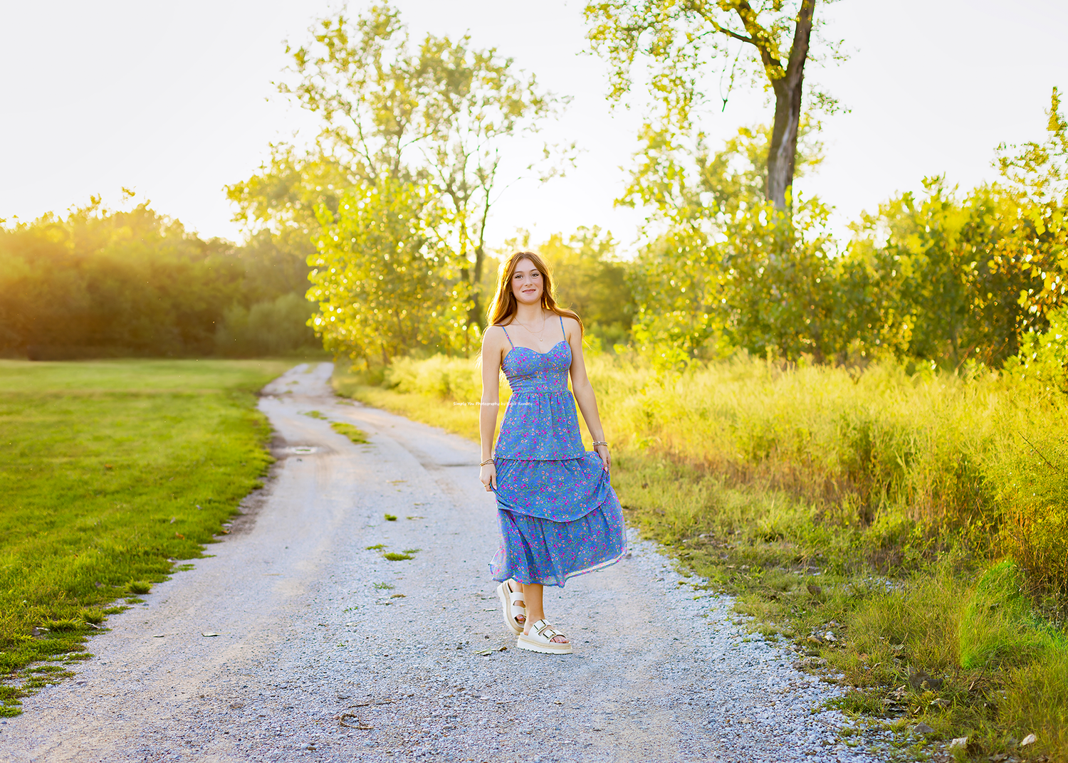 Young woman in blue floral sundress walking on a gravel path surrounded by lush green trees at golden hour.