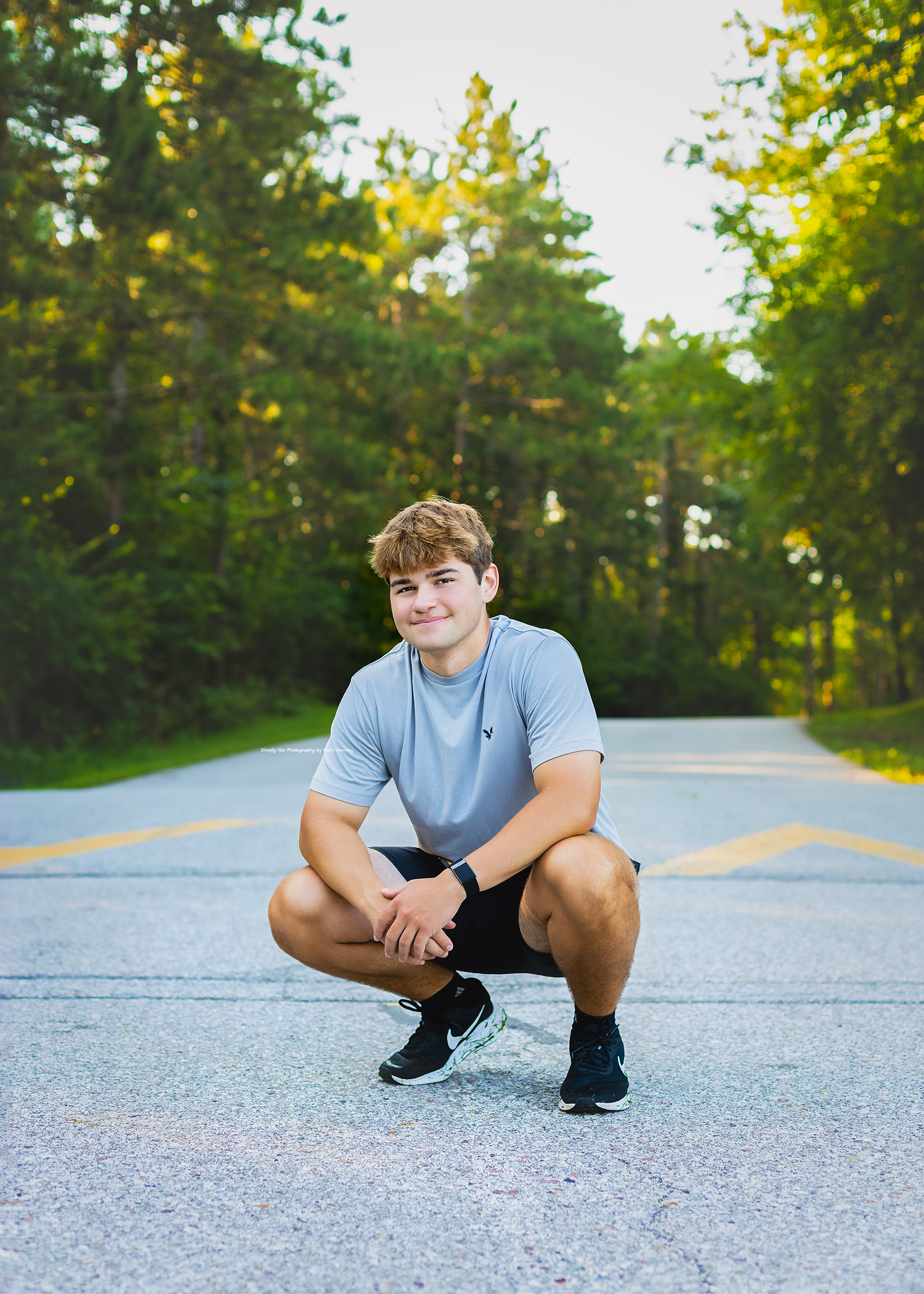Young man in gray shirt and black shorts crouching on a tree-lined road, smiling confidently outdoors.