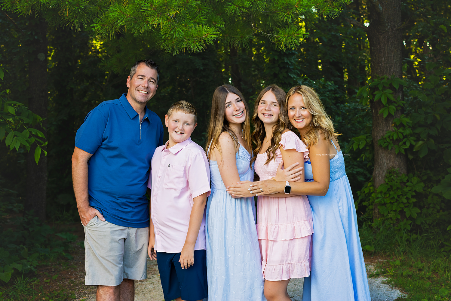 Happy family of five posing outdoors among lush green trees, wearing coordinated blue and pink outfits.