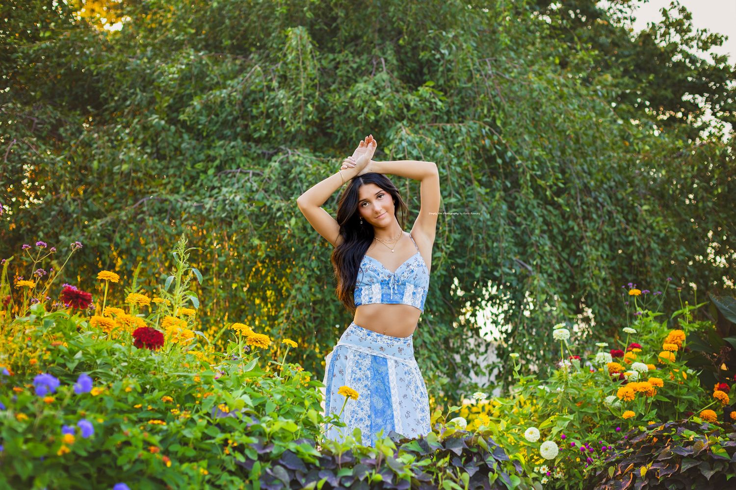 Young woman in blue floral two-piece outfit posing gracefully among colorful garden flowers at golden hour.