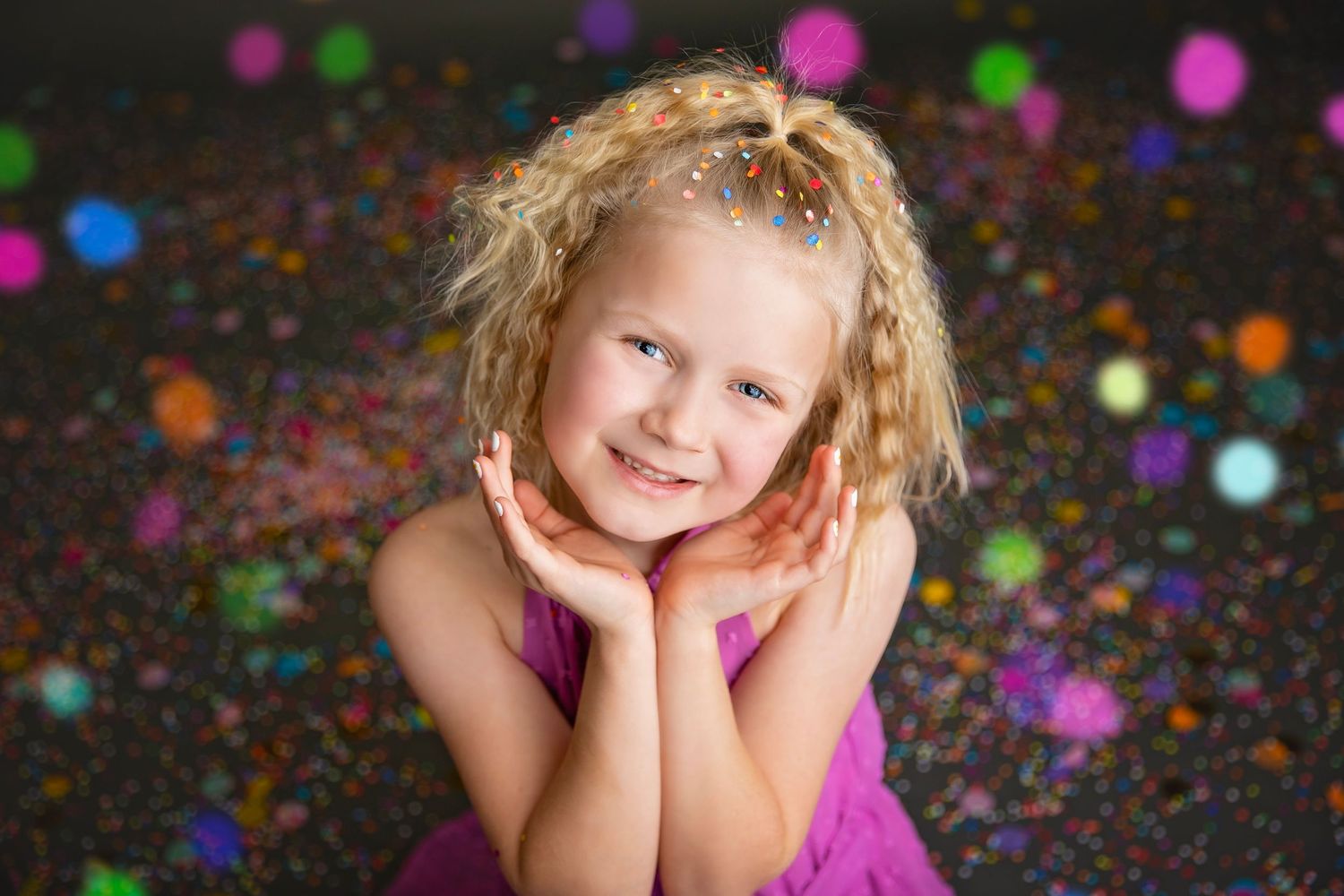 A cheerful blonde girl in a purple dress poses with hands under chin surrounded by colorful confetti bokeh lights.