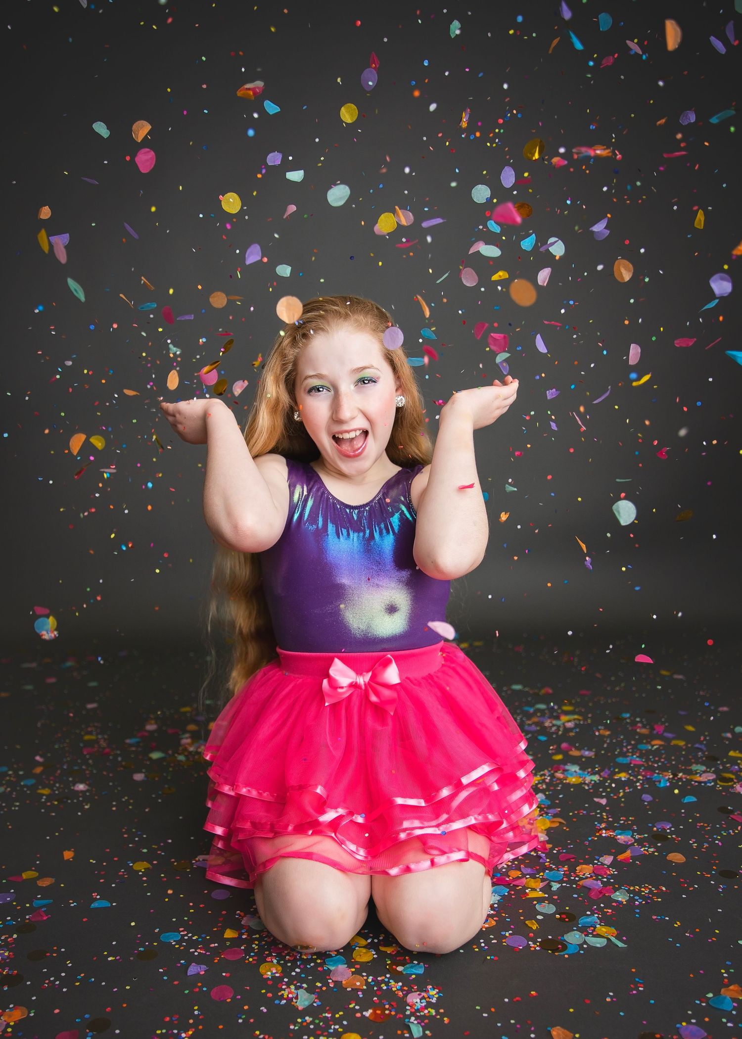 An excited young girl in a purple top and pink tutu kneels with hands raised as colorful confetti falls around her.