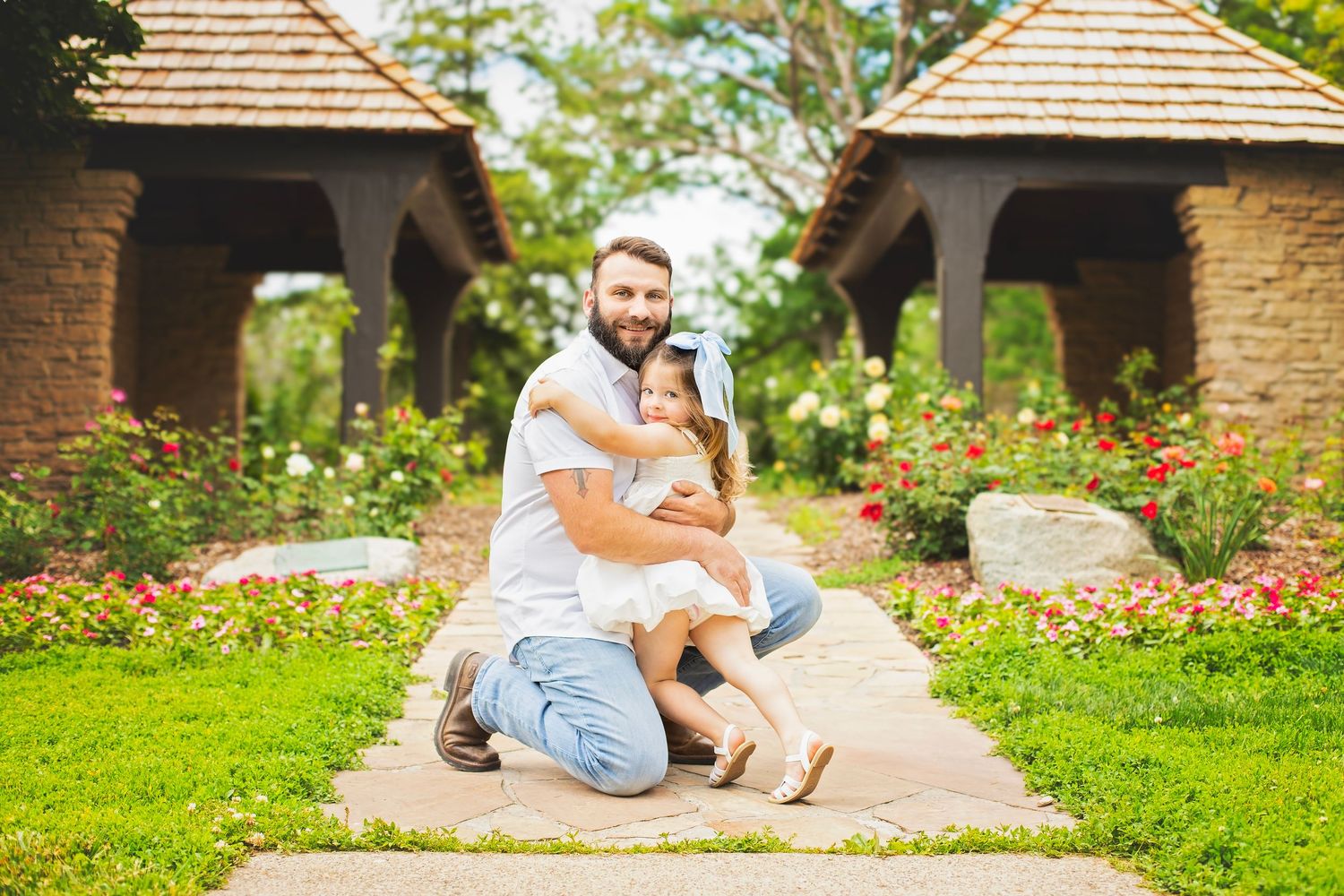 Loving father kneeling on garden path hugging his young daughter in white dress among colorful roses.
