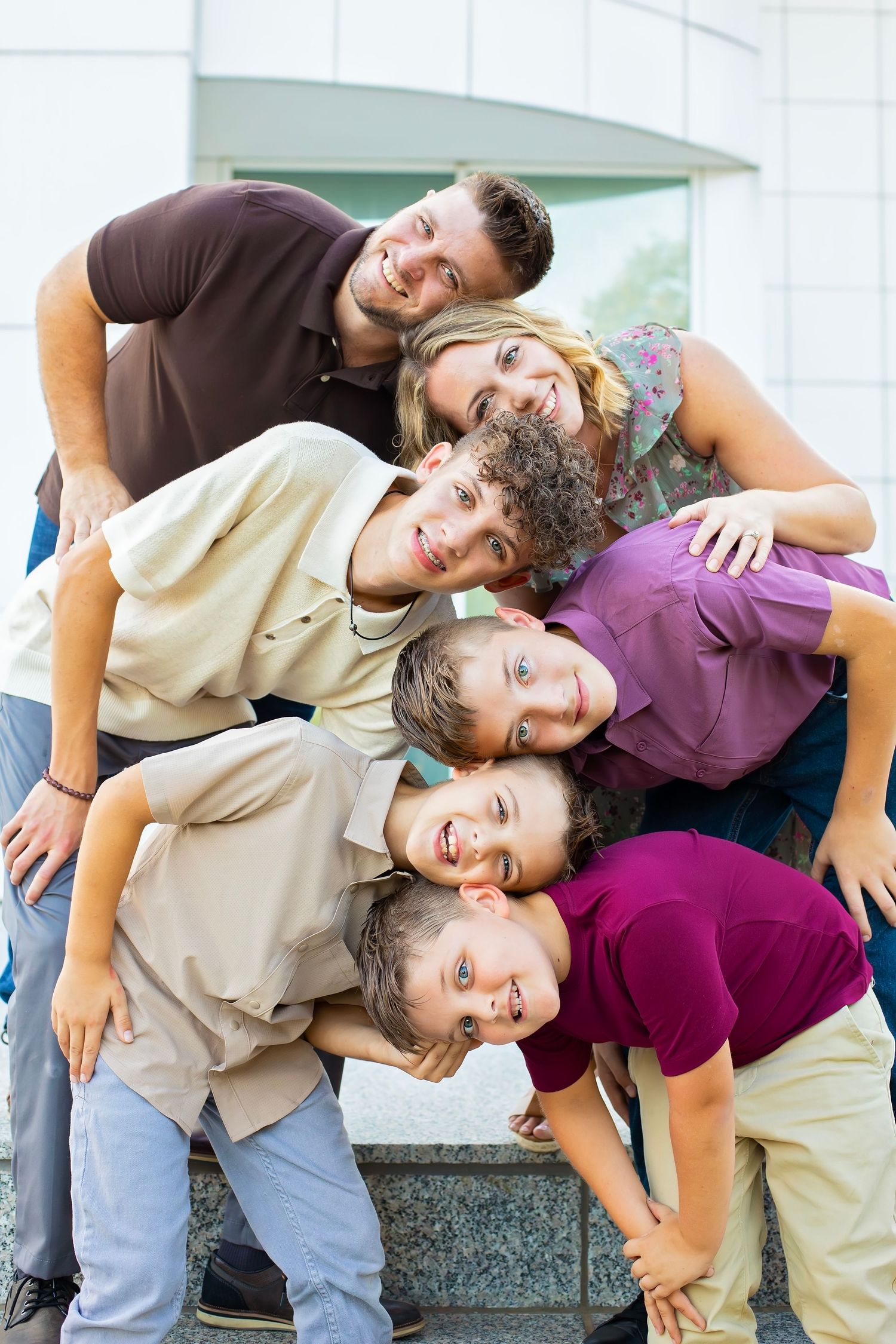 A joyful family of six stacks their heads together for a fun portrait outside a modern building.