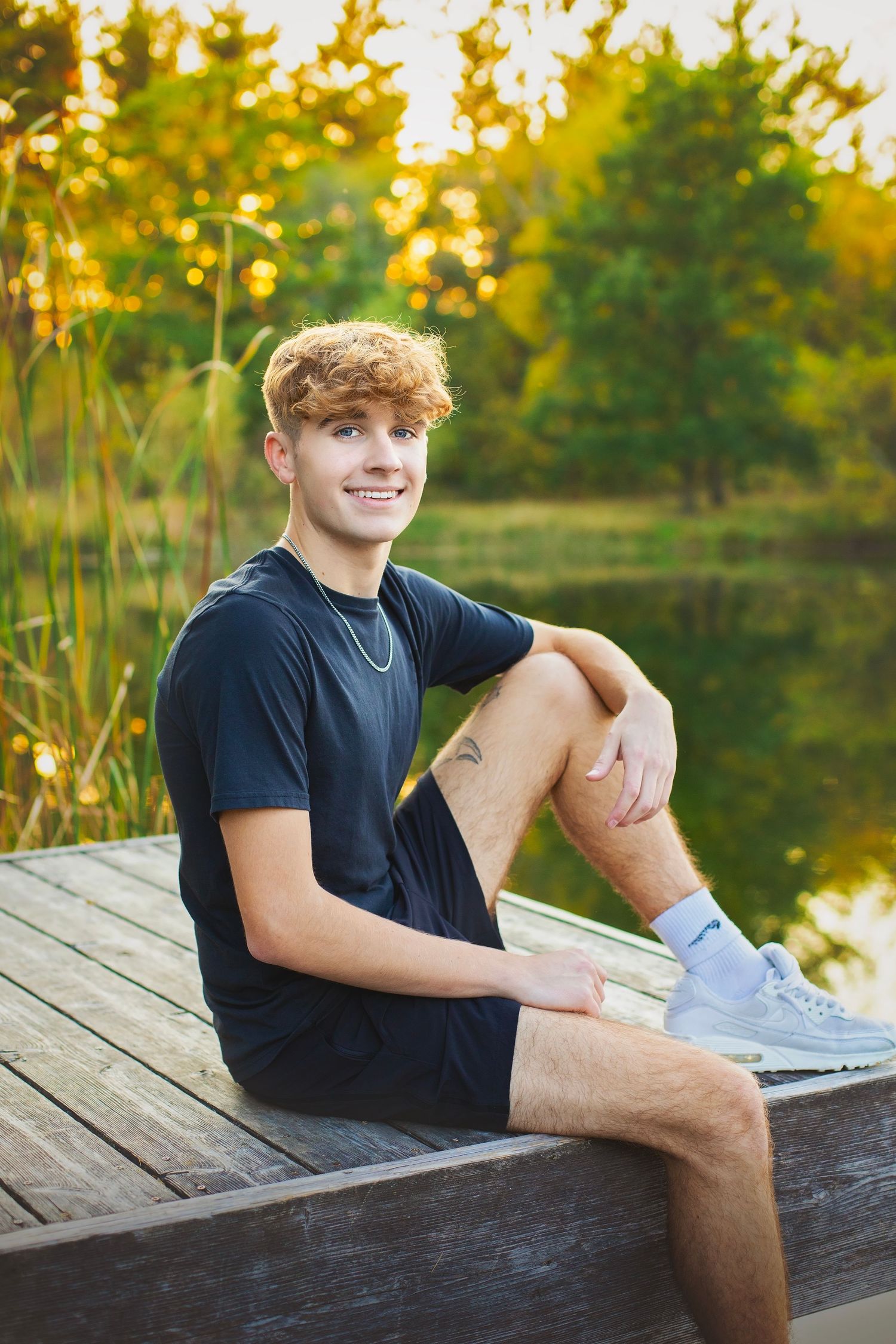 Curly redhead teen boy smiling on a wooden dock by a pond during golden hour senior photo session.